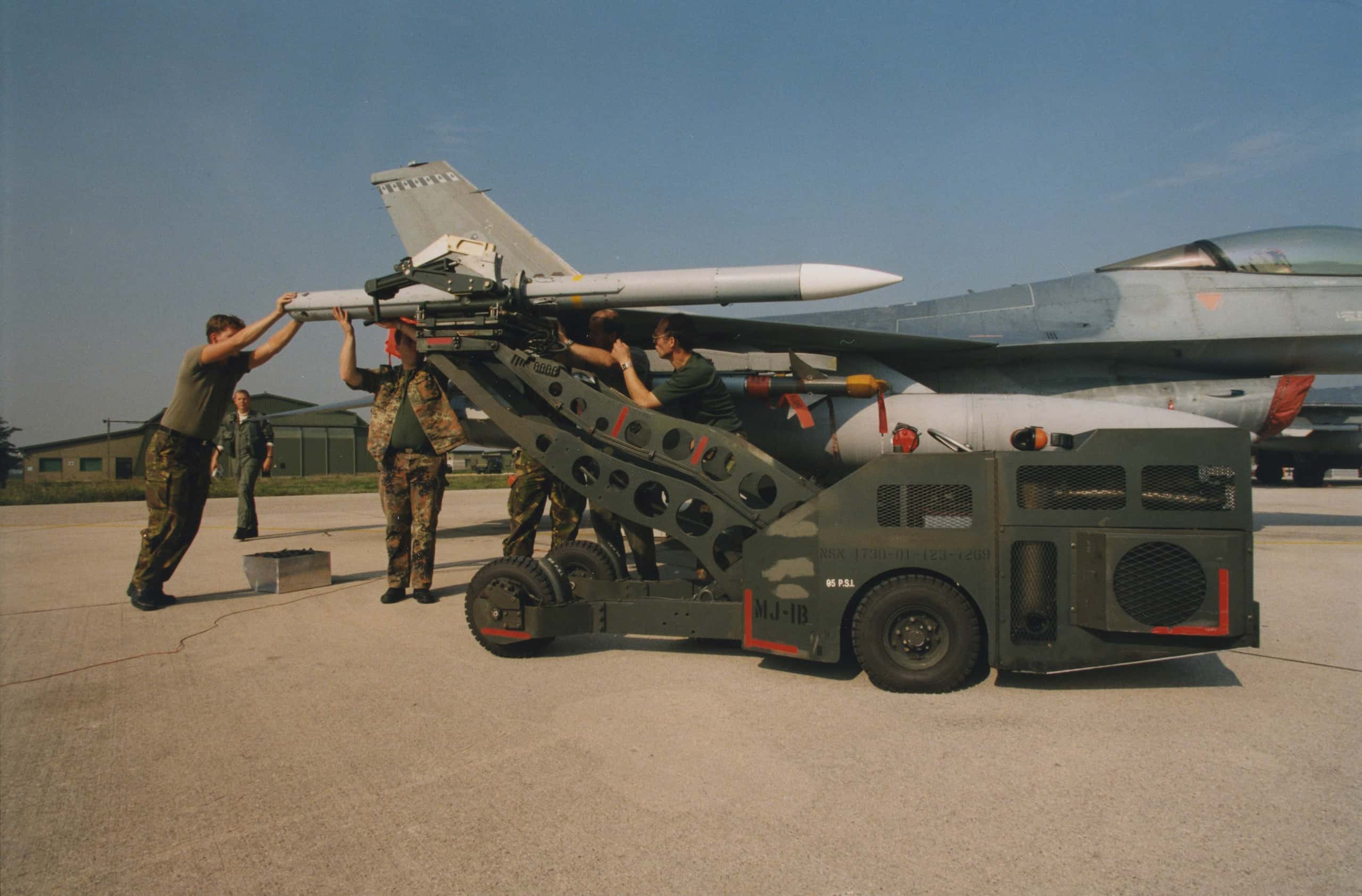 Kleurenfoto. Vijf militairen, sommigen half verscholen, werken aan een raket die aan het uiteinde van een vliegtuigvleugel wordt gemonteerd. De raket wordt door een takelarm van een takelwagentje vastgehouden.