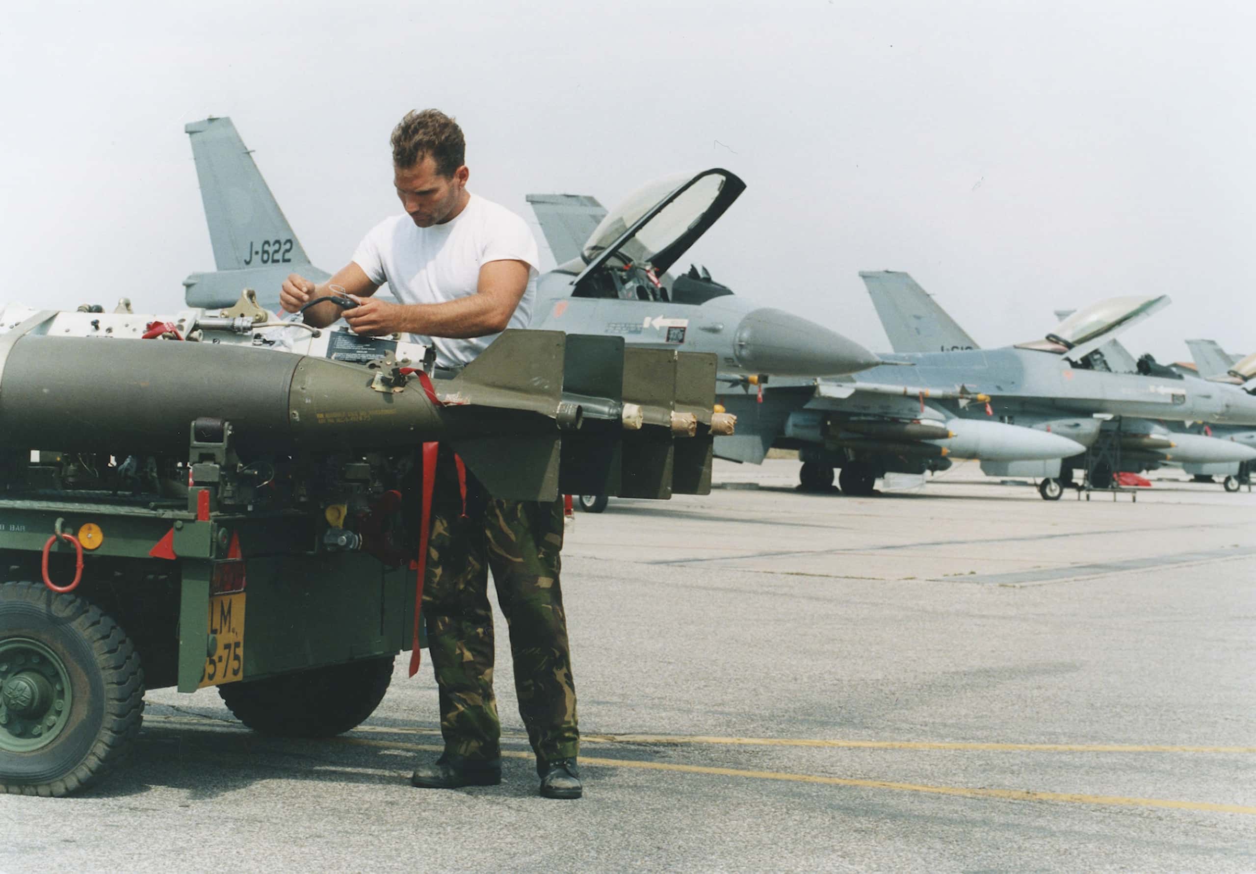 Kleurenfoto. Een militair in een wit t-shirt werkt aan drie bommen die op een aanhangwagen liggen. In de achtergrond staan meerdere gevechtsvliegtuigen op een rij.