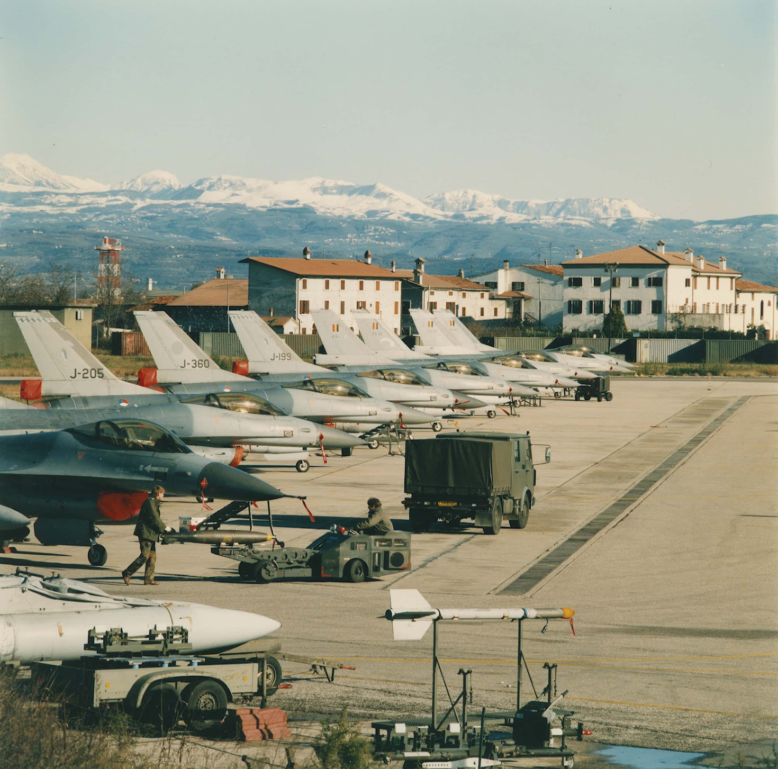 Kleurenfoto. Acht gevechtsvliegtuigen staan in de buitenlucht op een rij, de meest linker is slechts half te zien. De eerste drie hebben een letter en cijfercombinatie op de staart, de laatste vier het zwart-geel-rood van de Belgische vlag. Voor de vliegtuigen rijdt een vrachtwagen en zijn twee militairen aan het werk. In de achtergrond huizen en bergen.