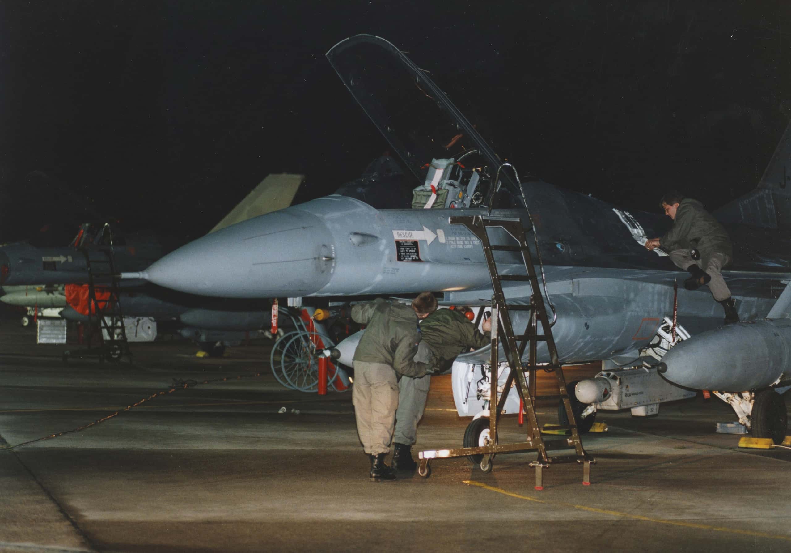 Kleurenfoto, ’s avonds laat of ’s nachts genomen. Drie militairen voeren werkzaamheden uit aan een gevechtsvliegtuig. Rechts zit één van hen op een vleugel, de andere twee staan aan de voorkant van het toestel. De cabine staat open en er staat een trappetje naast.