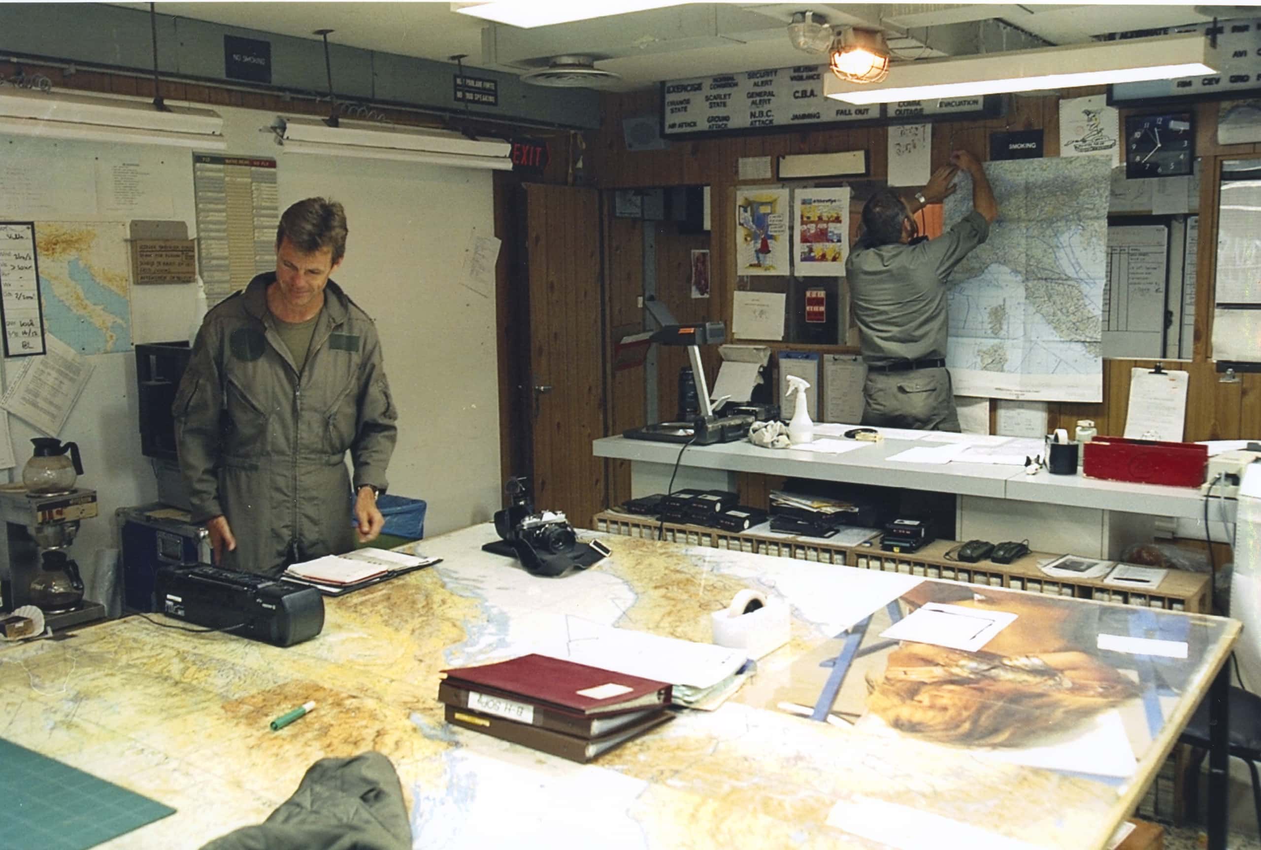 Kleurenfoto. Twee militairen staan in een kantoorruimte, de linker draagt een overall. Op de tafel in het midden liggen kaarten, mappen en andere bureauartikelen Ook aan de muren hangen veel kaarten.