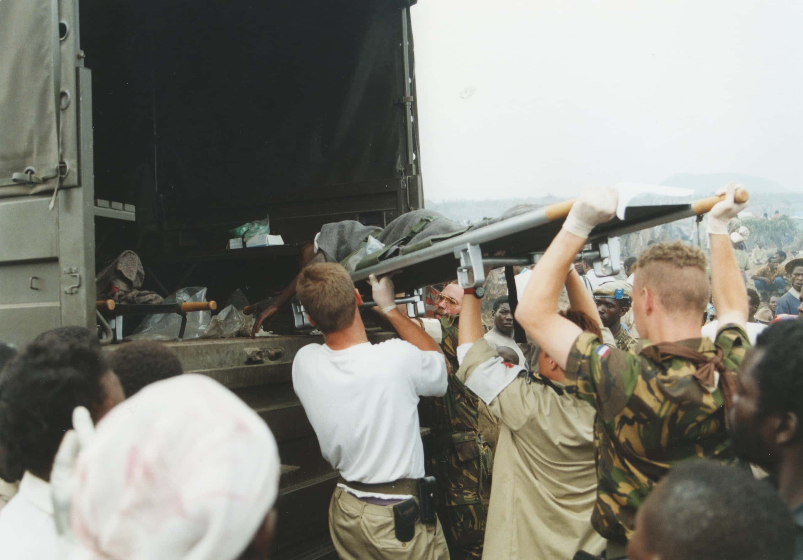 Kleurenfoto, vier mannen tillen boven hun hoofd een brancard met patiënt een vrachtwagen in. In de vrachtwagen ligt al een brancard. Twee mannen dragen groene camouflagekleding, eentje een beige shirt en een witte armband met een rood kruis, de vierde een wit T-shirt. Om hen heen staan Afrikaanse vluchtelingen tot te kijken.