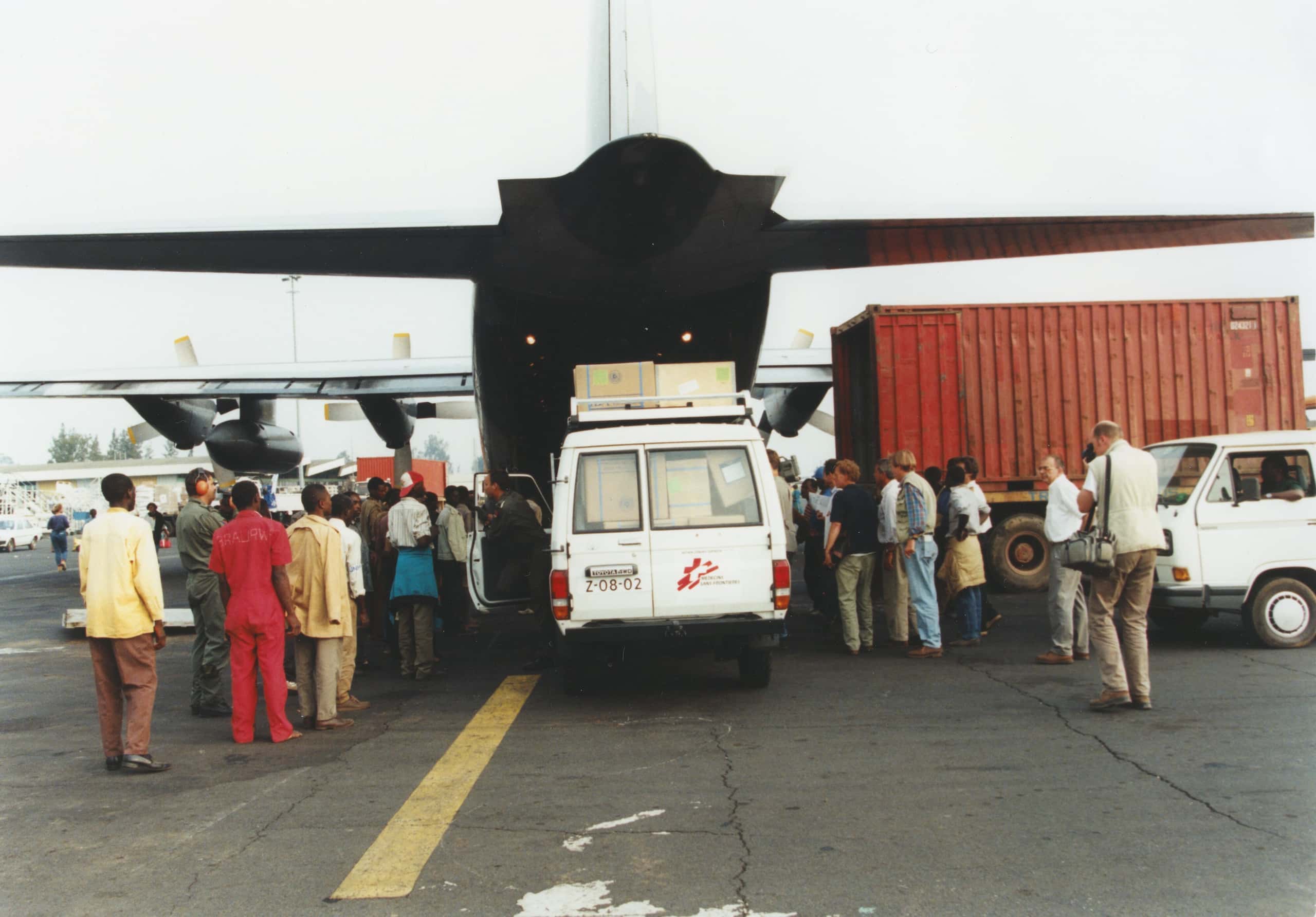 Kleurenfoto, bij de achterkant van een militair vliegtuig staat een grote groepen mensen te kijken hoe vanaf de achterkant geziene een witte bestelwagen naar buiten komt. De bestelwagen is volgeladen met dozen, iemand stapt aan de linkerkant uit. Rechts naast het vliegtuig een rode container en een wit busje.