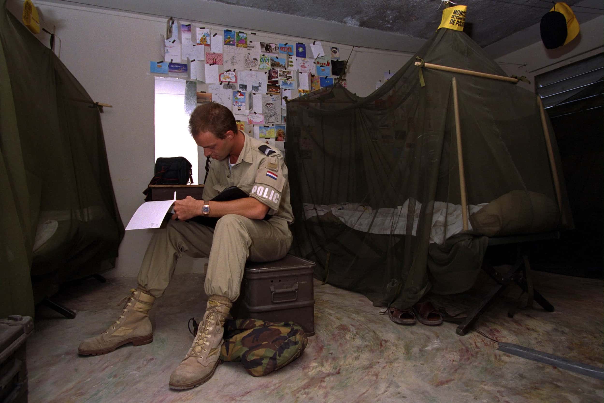 Kleurenfoto, binnen in een slaapkamer. Een militair in beige kleding zit op een kist, rechts een bed met een donkere klamboe erover. De militair schrijft iets op een blocnote, er is één vel papier omgeslagen. Aan de muur, naast een smal raam, hangen tientallen brieven, kaarten en foto’s.