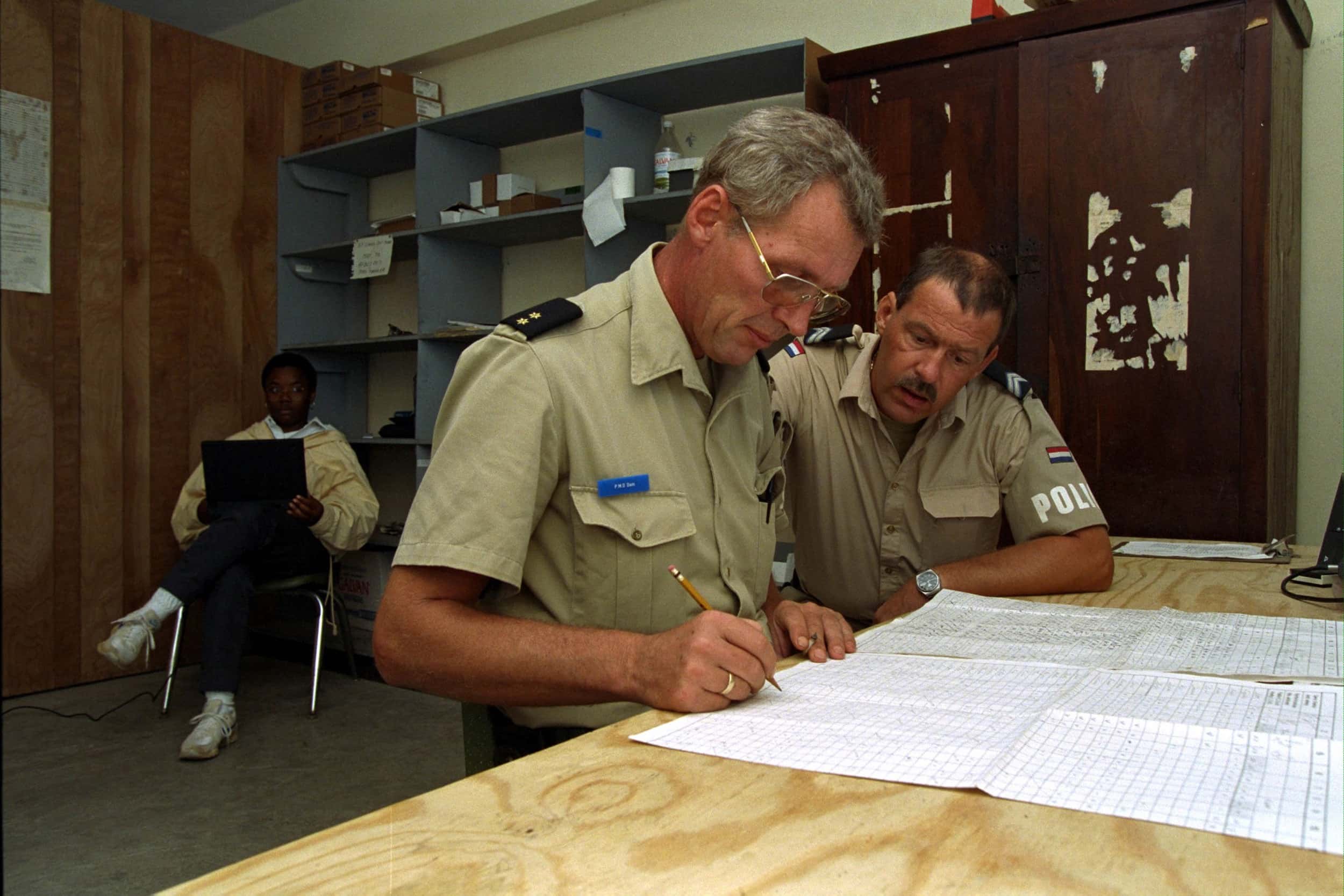Kleurenfoto, binnen in een kantoorruimte. Twee militairen in beige kleding buigen zich over twee grote vellen papier dat op een tafel ligt. De linker draagt een bril en heeft een potlood vast. Op de achtergrond zit iemand op een stoel iets te lezen, tegen de muur staat een stellingkast met kantoorartikelen.