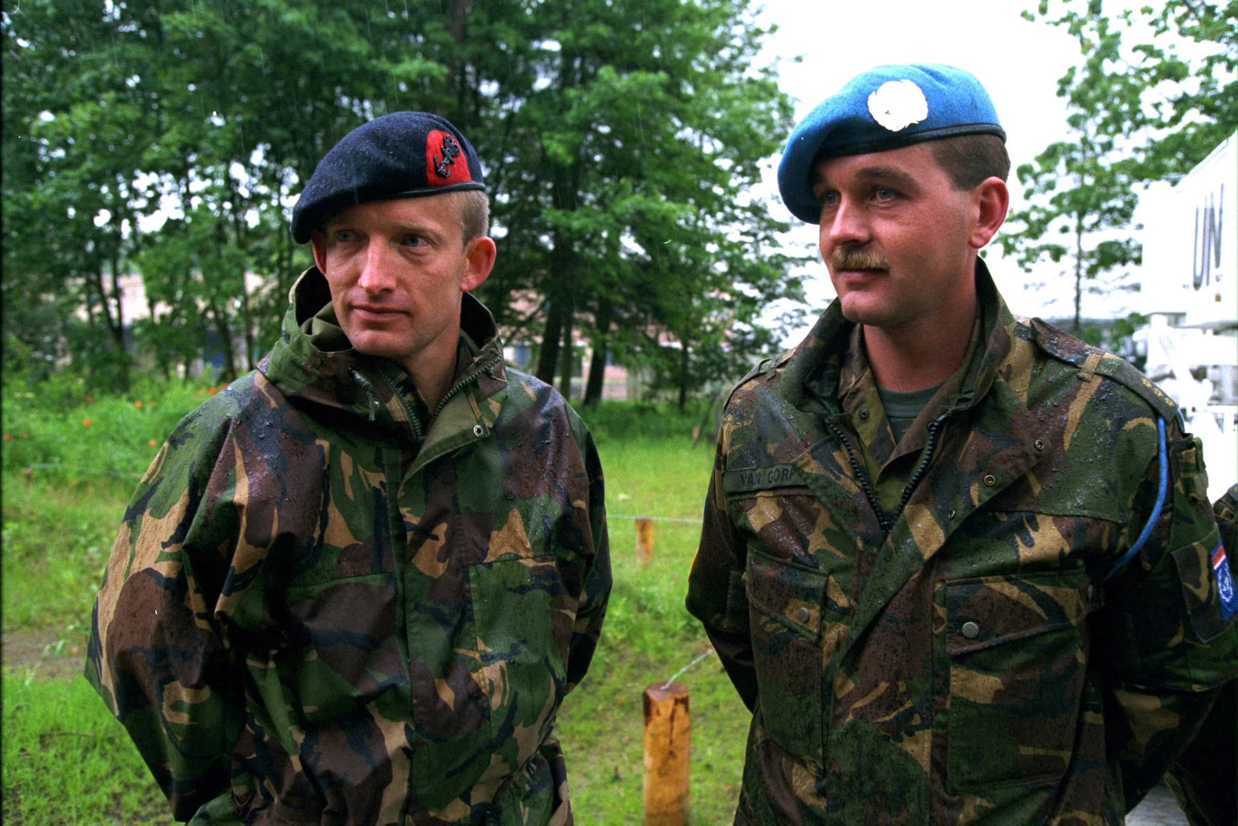 Kleurenfoto, buiten. Twee militairen in groene camouflagekleding kijken naar iets of iemand links buiten de foto, beide hebben hun handen op de rug. De rechter militair draagt een lichtblauwe baret, de linker een donkerblauwe baret. Hun kleding is nat van de regen. Op de achtergrond een grasveldje, bomen en rechts een wit voertuig met de letters UN.