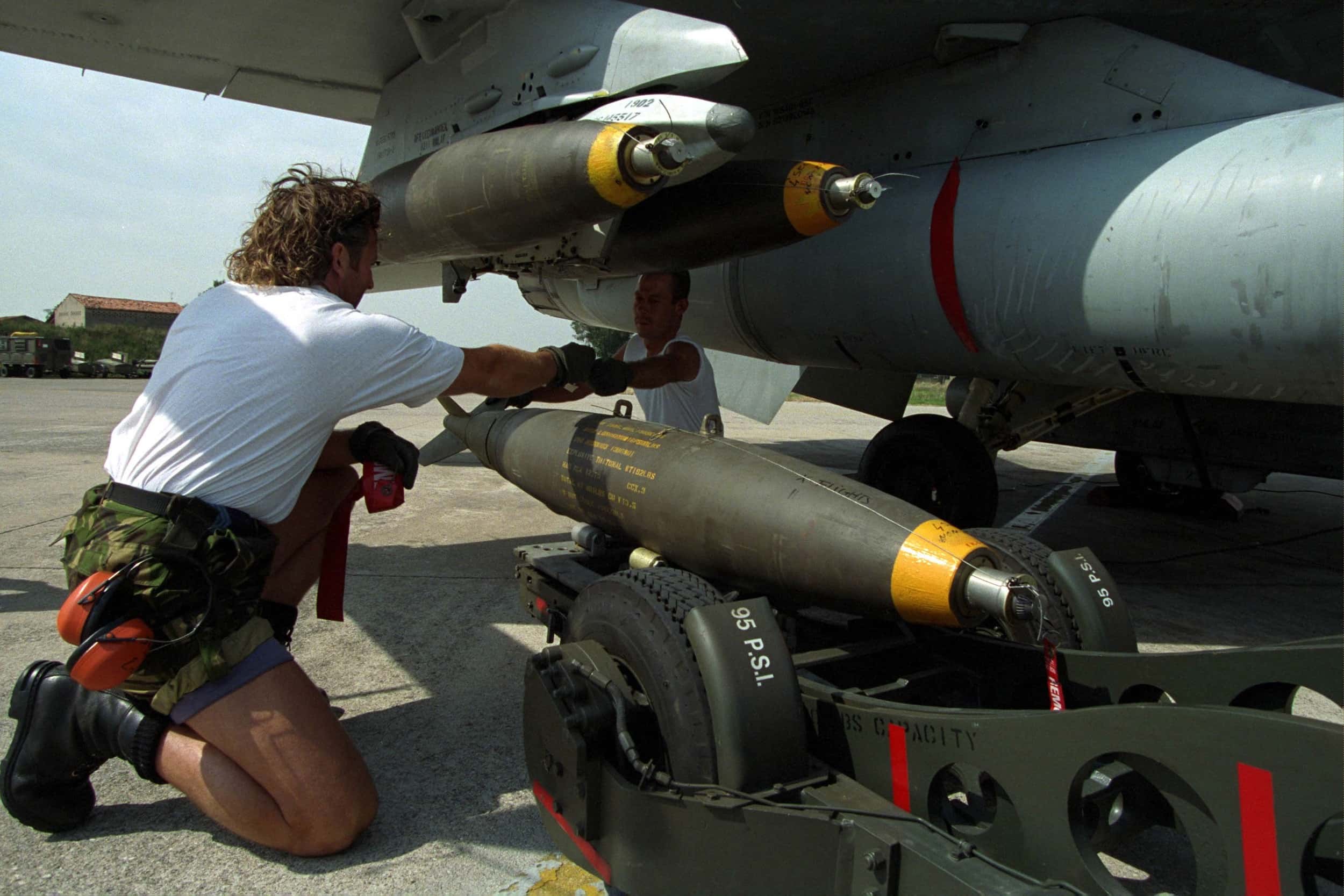 Kleurenfoto, buiten op een vliegveld. Twee mannen zitten onder de vleugel van een militair vliegtuig, de man links zit op één knie. Tussen hen in drie bommen, die zij aan de vleugel boven hun hoofd vastmaken. Zij reiken iets naar elkaar aan. Aan de korte broek van de man links hangt een koptelefoon.