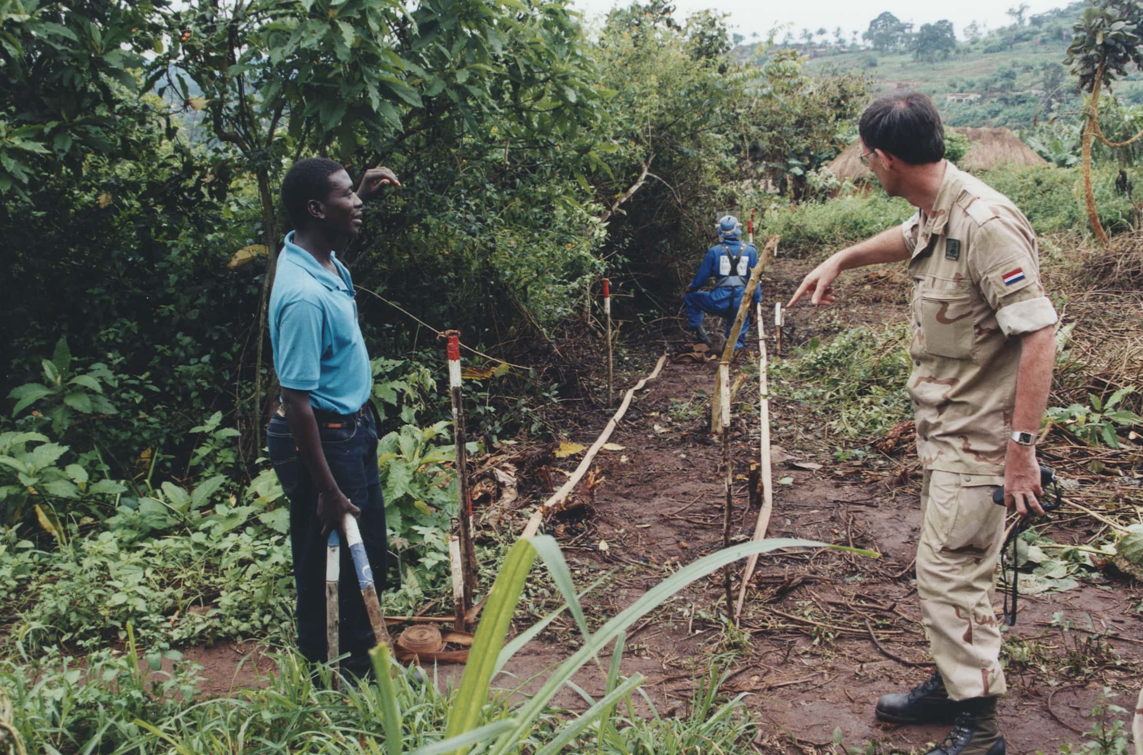 Kleurenfoto. Drie mannen zijn in het struikgewas aan het werk. Rechts staat een militair in uniform in camouflagepatroon, hij praat met de man links die burgerkleding draagt. Verderop zit de derde man, gekleed in blauwe overall op één knie met zijn rug naar de camera. Deze man draagt ook een vizier voor zijn gezicht en een bombestendig vest. Tussen de man in burger en de man in overall zijn drie houten paaltjes met rood-witte bovenkant in de grond geslagen. De rechterkant van het struikgewas is gekapt, de linkerkant niet. In de achtergrond een heuvelachtig grasland met veel bomen en struiken.