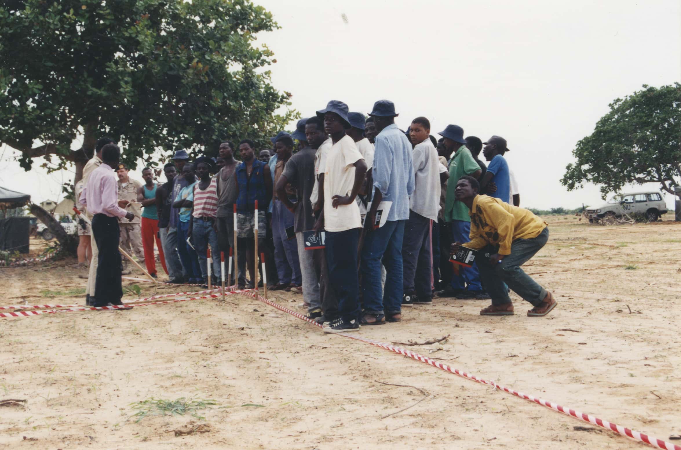 Kleurenfoto. Een grote groep mannen staat op een kleine zandvlakte dicht op elkaar. Ze dragen burgerkleding, velen ook een zonnehoed en het merendeel kijkt in de camera. Sommigen hebben een schrift in hun handen. Voor de groep zijn zes houten paaltjes met rood-witte bovenkant in een clustertje in de grond geslagen, op de grond lopen meerdere rood-witte veiligheidslinten. Voor de groep staan twee mannen, één in burgerkleding en de ander in militair uniform. Een andere militair, ook in uniform, staat naast de groep te luisteren. In de achtergrond een paar bomen, waar auto’s onder staan.