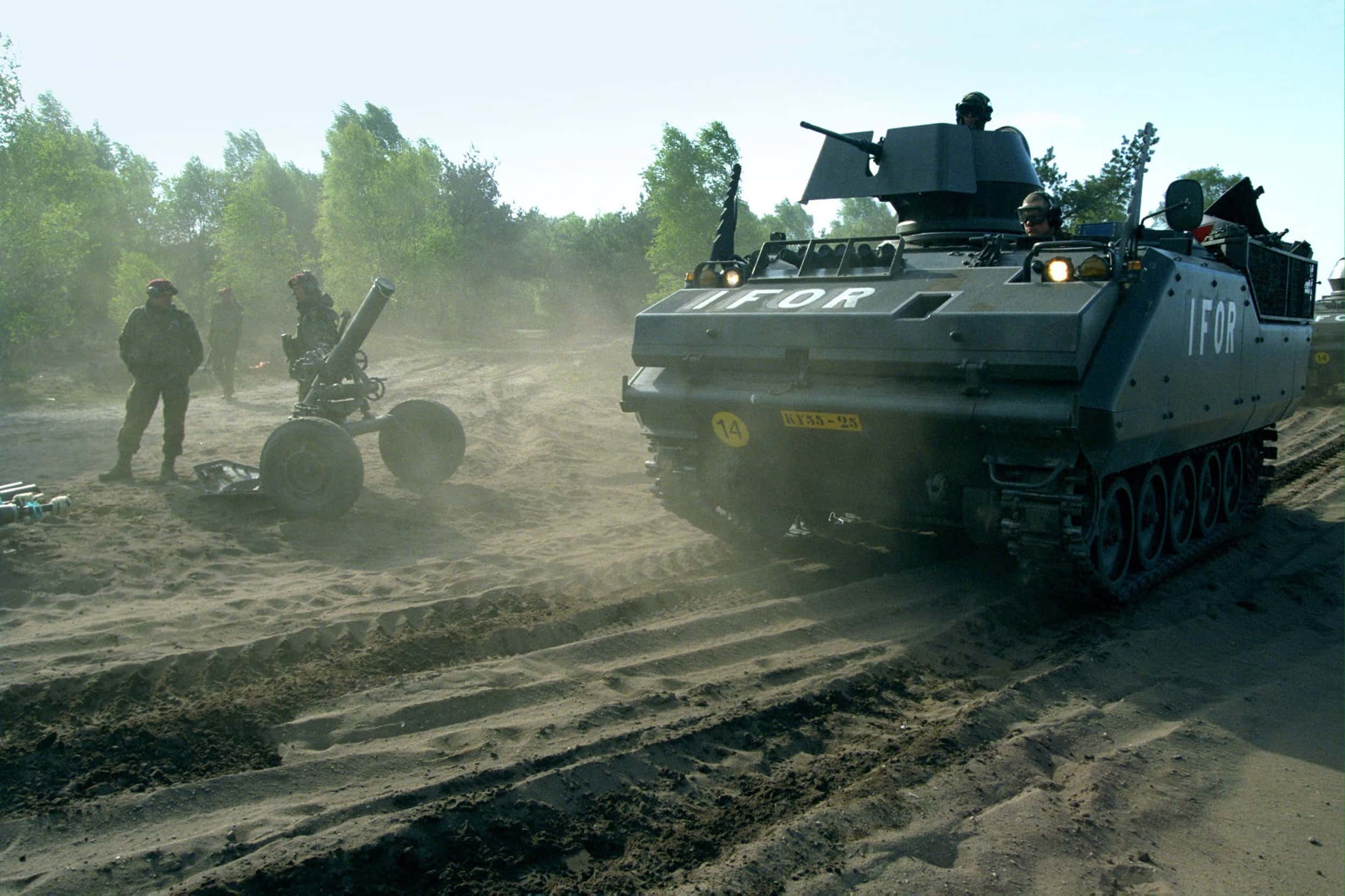 Kleurenfoto. Een grote pantserwagen rijdt op een zandvlakte, op de voor- en zijkant staat in witte letters IFOR geschreven. Uit twee openingen aan de bovenkant steken twee militairen naar buiten. Bovenop is een geweer met beschermpaneel geplaatst. Links op de grond staat een militair met naast zich een mortier op twee dikke wielen. In de achtergrond bomen en struiken.
