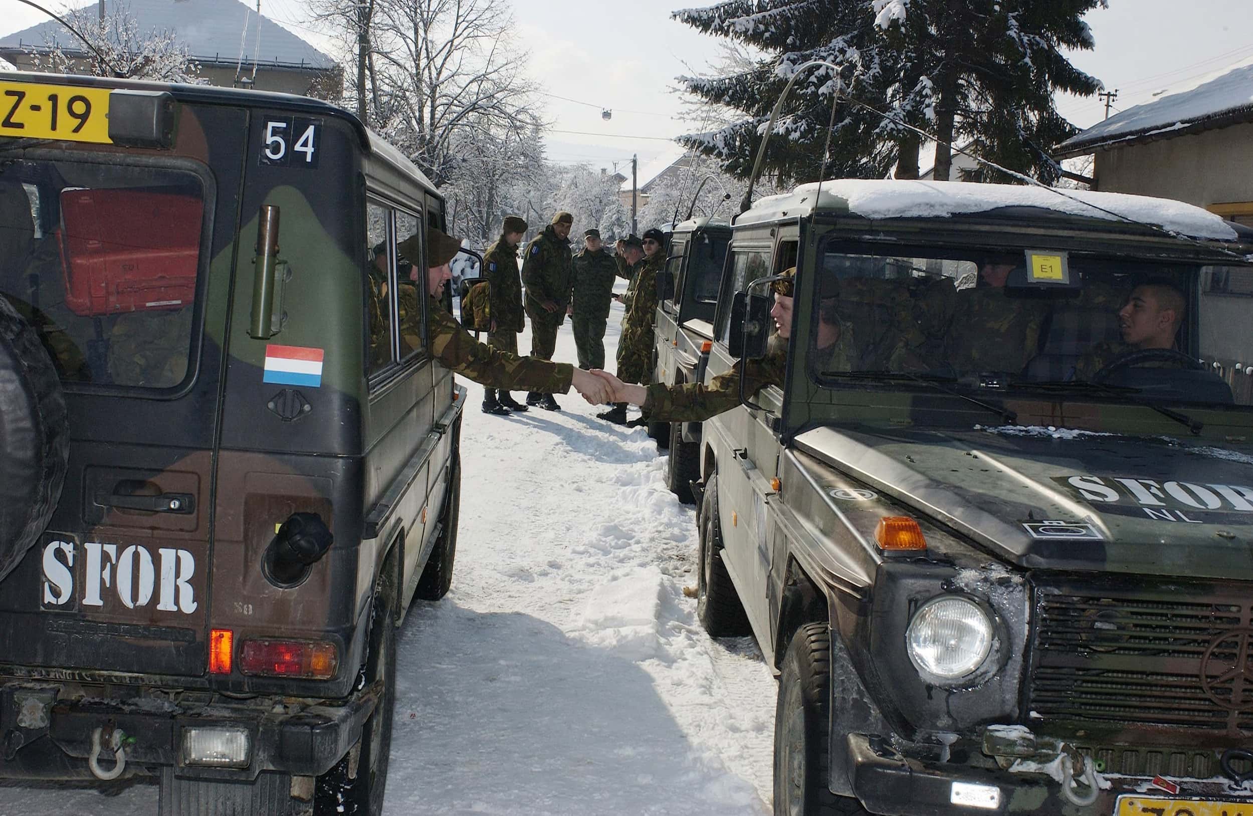 Kleurenfoto. Twee jeeps staan naast elkaar geparkeerd, de linker met de achterkant naar de camera, de rechter met de neus naar de camera. Uit beide jeeps steekt een militair naar buiten die de hand schudt van de ander. Op de motorkap van de rechter jeep en achterop de linker jeep staat in het wit SFOR geschreven. In de achtergrond staat een groepje militairen in camouflage-uniform ernaar te kijken. Op de grond en in de bomen op de achtergrond ligt sneeuw.