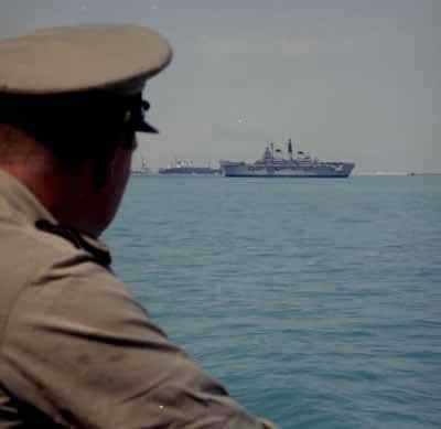 Kleurenfoto, buiten op het dek van een marineschip. Een man in beige kleding en een beige platte pet op kijkt over het water naar drie marineschepen die in de verte liggen. Het water beneden is blauw, de lucht boven de schepen is grijs.