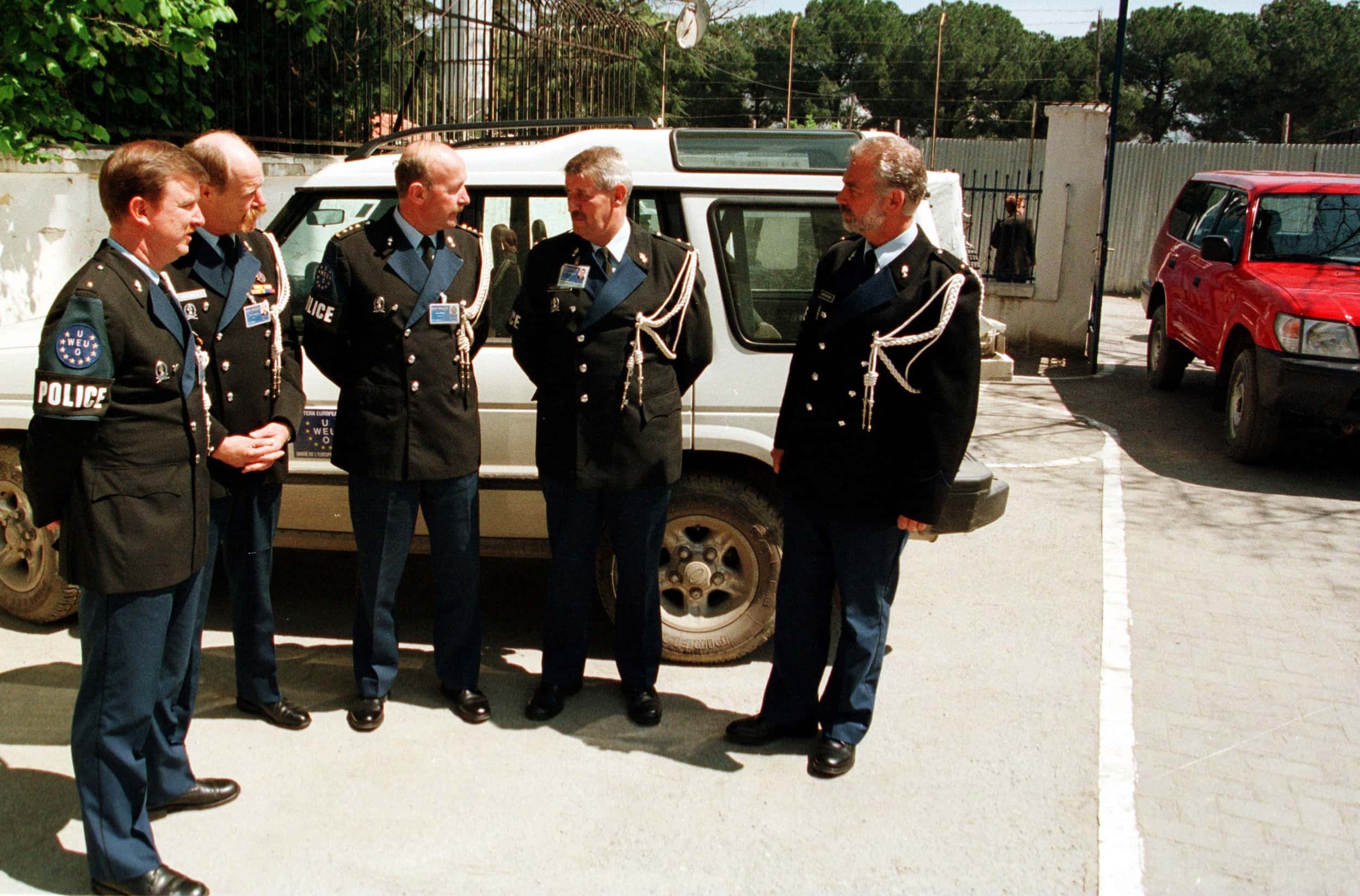 Kleurenfoto, buiten op een parkeerplaats. Vijf mannen zijn met elkaar in gesprek, zij dragen allemaal hetzelfde donkerblauwe pak en witte koorden om hun linkerschouder. Meteen achter hen een wit voertuig, rechts komt een rode auto de parkeerplaats op gereden. Op de achtergrond achtereenvolgens een hek dat overgaat in een muur, de straat en een ander hek.