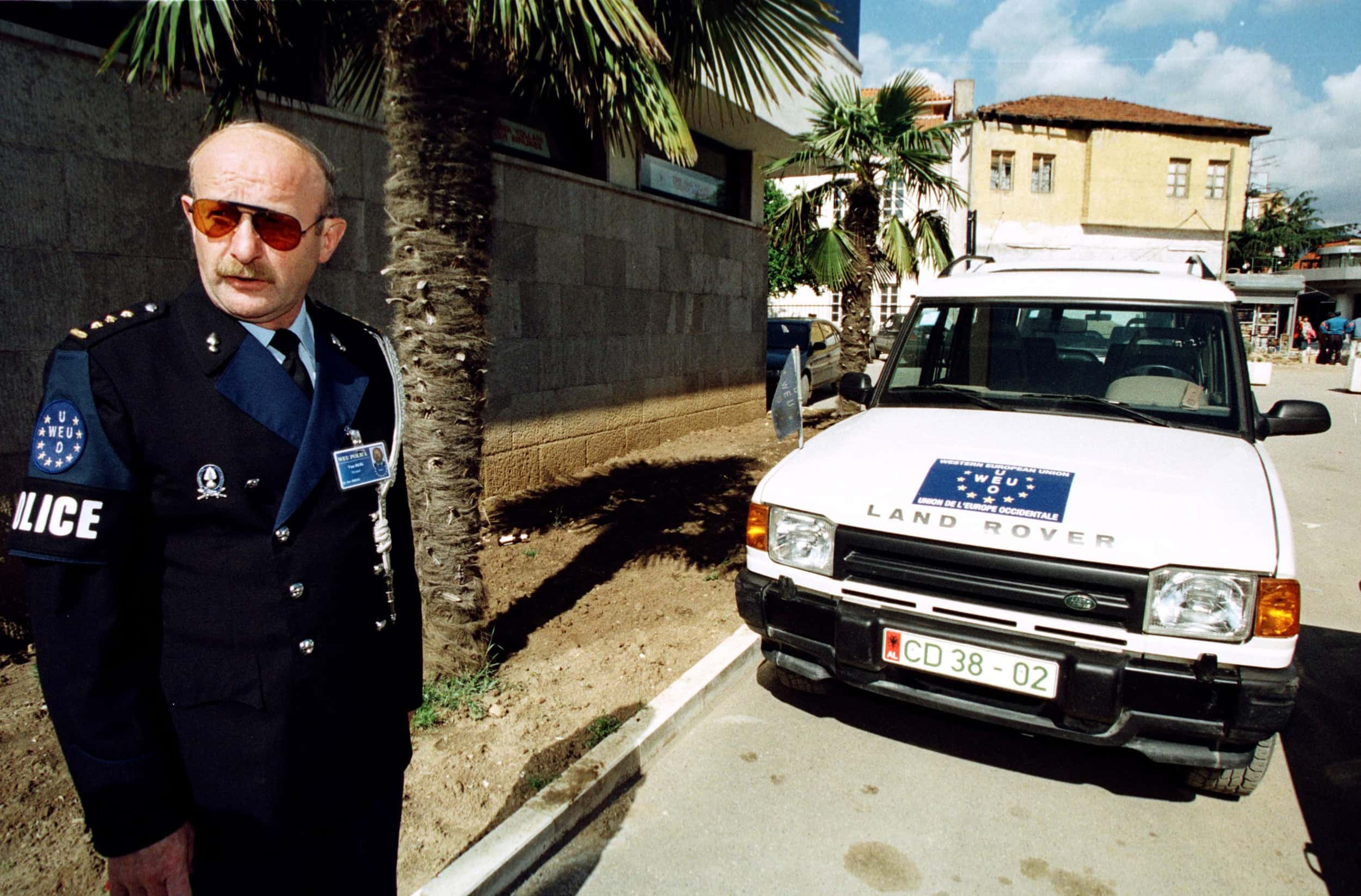 Kleurenfoto, buiten langs de kant van de weg. Een man met snor en zonnebril kijkt naar links buiten beeld, hij draagt een donker pak met een wit koord om zijn linkerschouder. Rechts van hem een wit voertuig met het woord Landrover en het logo van de operatie op de motorkap. In de berm op de achtergrond twee palmbomen en daarachter huizen.