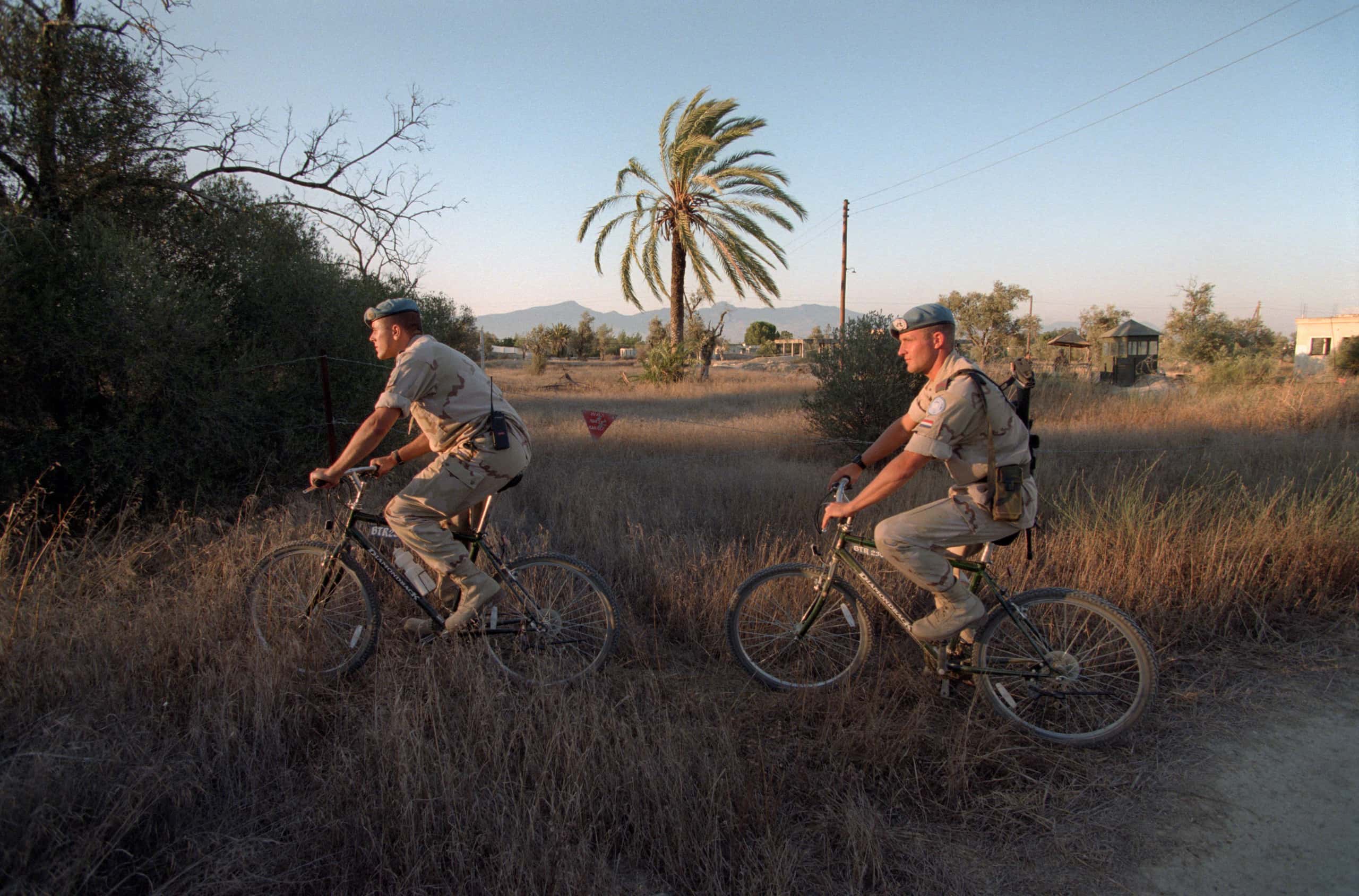 Kleurenfoto, twee militairen in camouflagekleding rijden achter elkaar op mountainbikes door het gras. Links is een heg, op de achtergrond een grote palmboom, een elektriciteitspaal en uitkijkposten.