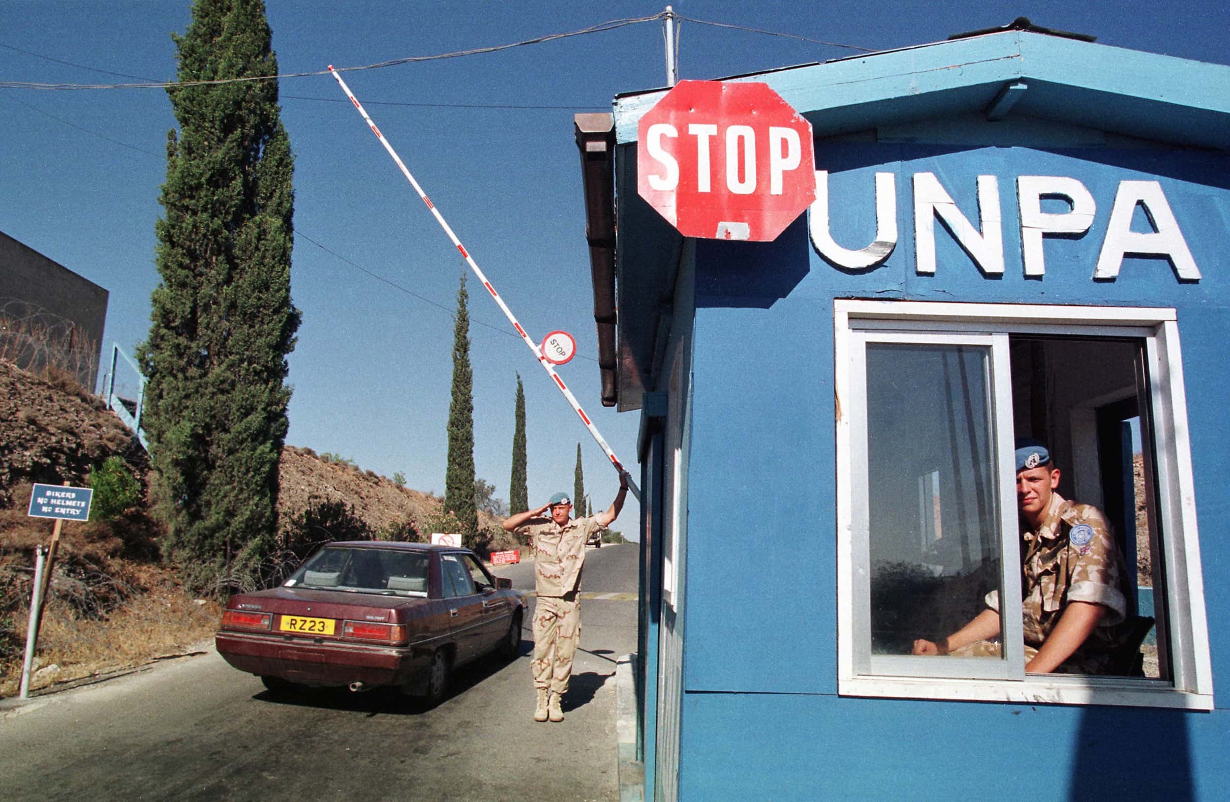 Kleurenfoto, rechts een blauw huisje met de letters UNPA en een stopbord, links daarvan een rood-witte slagboom en een rode auto. In het huisje zit een militair, terwijl een andere militair de slagboom omhoog houdt en een groet brengt. Links langs de weg een dijkje met cipressen.
