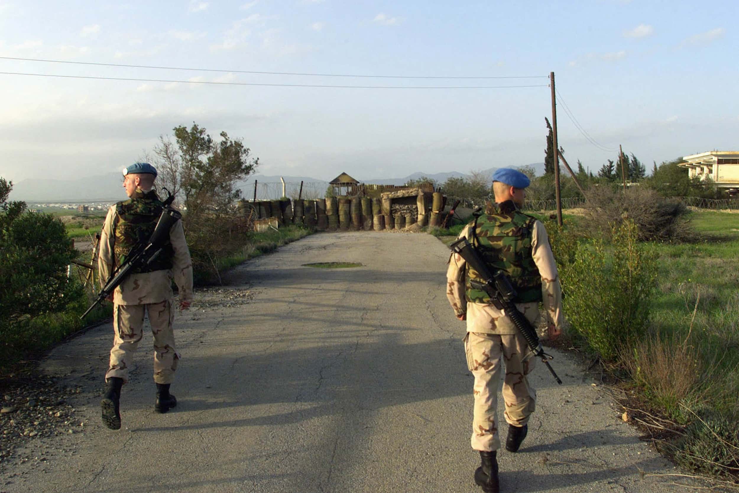 Kleurenfoto, twee militairen met geweren vanaf de rug gezien lopen op een weg, de één kijkt links, de ander naar rechts. Voor hen is een barricade, een hek met prikkeldraad en een uitkijktoren. Links en rechts van de weg is een berm met lage struiken, in de verre achtergrond bergen.