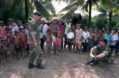Kleurenfoto, buiten. Rechts zit een militair in camouflagekleding in kleermakerszit op de grond, zijn armen zijn om z’n knieën geslagen. Links komt een andere militair, ook in camouflagekleding, naar hem toe gelopen. Achter hen een lange rij van lokale mannen en kinderen in burgerkleding. Daarachter palmbomen en het dak van een huis of hut.