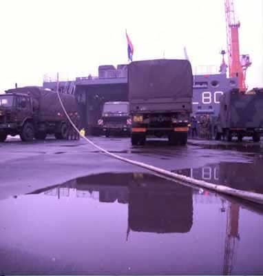 Kleurenfoto, buiten op een kade. Vier legervrachtwagens staan op een kade klaar om ingeladen te worden op een schip, dat achter de voertuigen zichtbaar is. Op de kade in de voorgrond een plas water, er loopt een kabel van de rechterbenedenhoek naar het dak van de meest linker vrachtwagen. Op het schip wappert een Nederlandse vlag, rechts in de achtergrond een hoge oranje hijskraan.