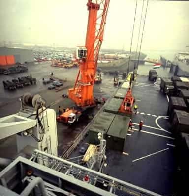 Kleurenfoto, van een hoogte genomen op een schip, uitkijkend over het schip en een kade. Een oranje hijskraan zet containers op het dek van een marineschip. Op het dek zijn meerdere containers, vrachtwagens en bemanningsleden te zien, aan het uiteinde wappert een Nederlandse vlag. Op de kade staan nog meer vrachtwagens. In de achtergrond nog een marineschip en de haventerrein.