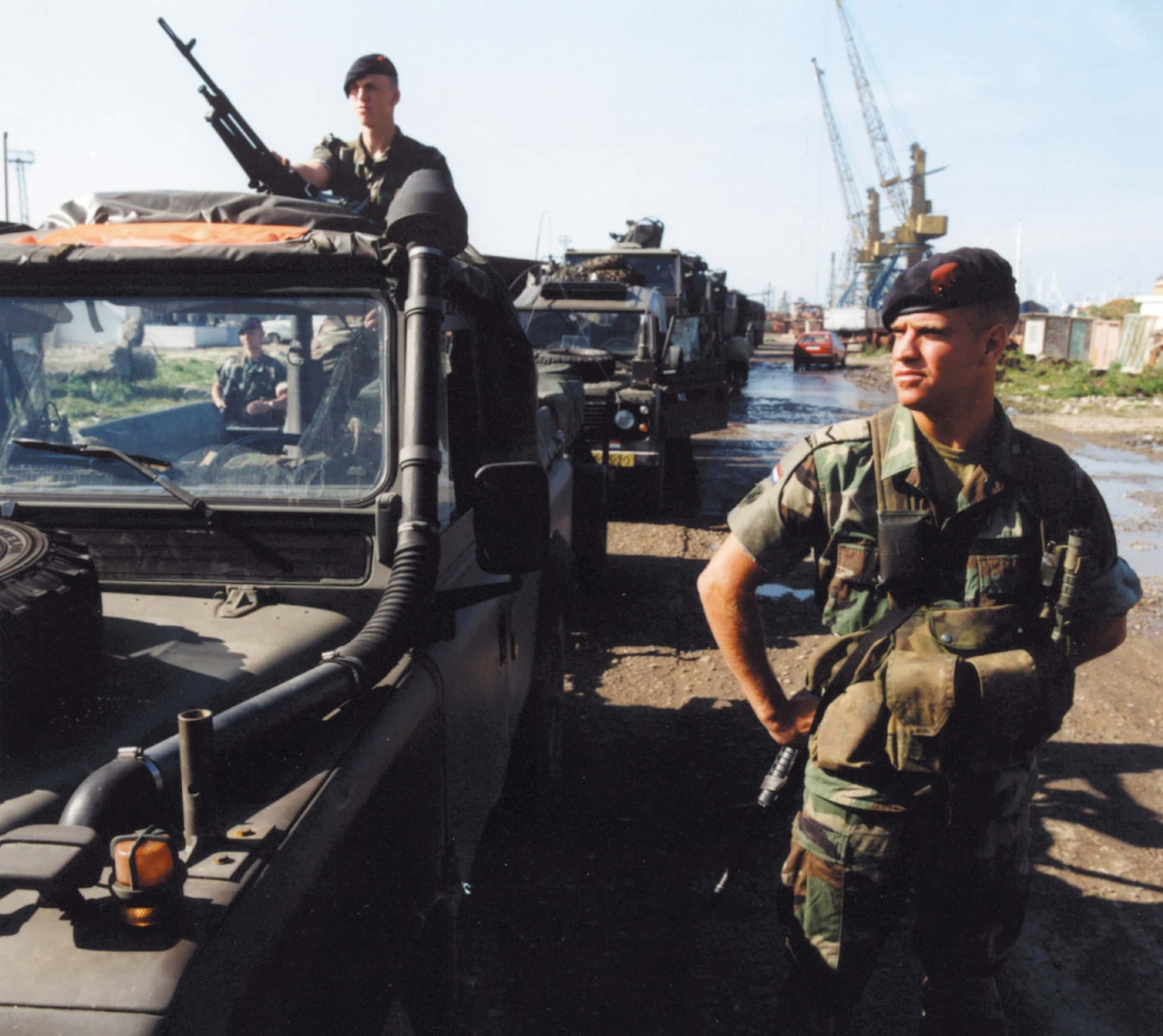 Kleurenfoto, buiten. Rechts staat een militair in camouflagekleding, hij draagt een baret en een geweer. Links een rij militaire voertuigen, bovenop de voorste staat een andere militair met een mitrailleur in de aanslag, hij steekt boven de cabine uit. Op de achtergrond, voorbij de rij voertuigen, een paar havenhijskranen en een rode auto.