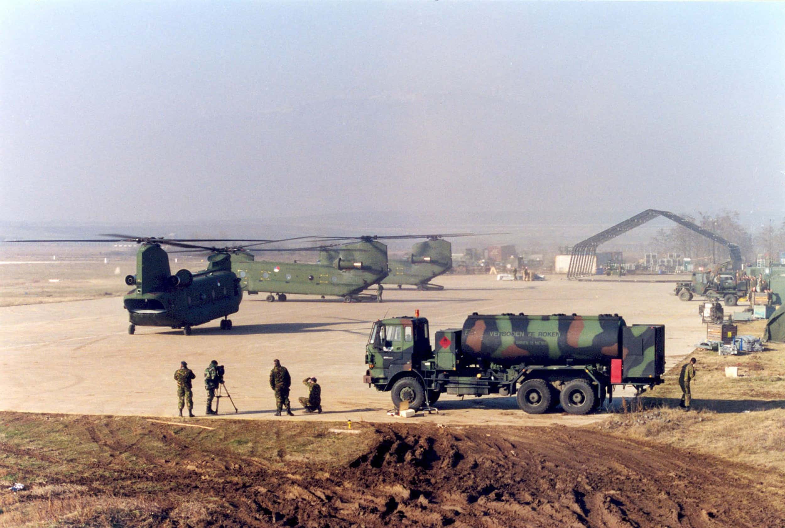 Kleurenfoto, buiten overkijkend over een vliegplatform. Op het platform staan drie helikopters met twee sets rotorbladen, rechts op de voorgrond een vrachtwagen in camouflagekleuren geschilderd. Links daarnaast staan vier militairen, één daarvan kijkt door een camera op een driepoot, de meest rechter zit op één knie. Achter de vrachtwagen staat nog een militair. In de achtergrond de installatie een hangar, zonder overkapping.