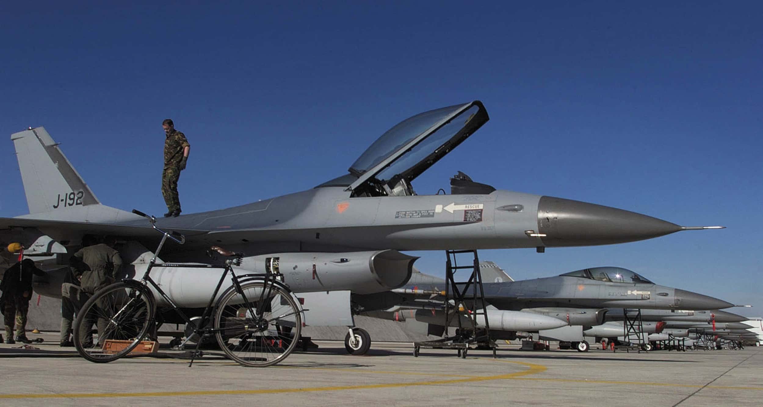 Kleurenfoto, buiten op een vliegveld. Een F-16 staat geparkeerd, de koepel van de cockpit staat open, daarachter staat een militair. Op de staart staat J-192. De militair bovenop het toestel praat met drie andere militairen op de grond. Links in de voorgrond een fiets, achter het toestel een lange rij F-16’s. De zon schijnt, er is geen wolkje aan de lucht.