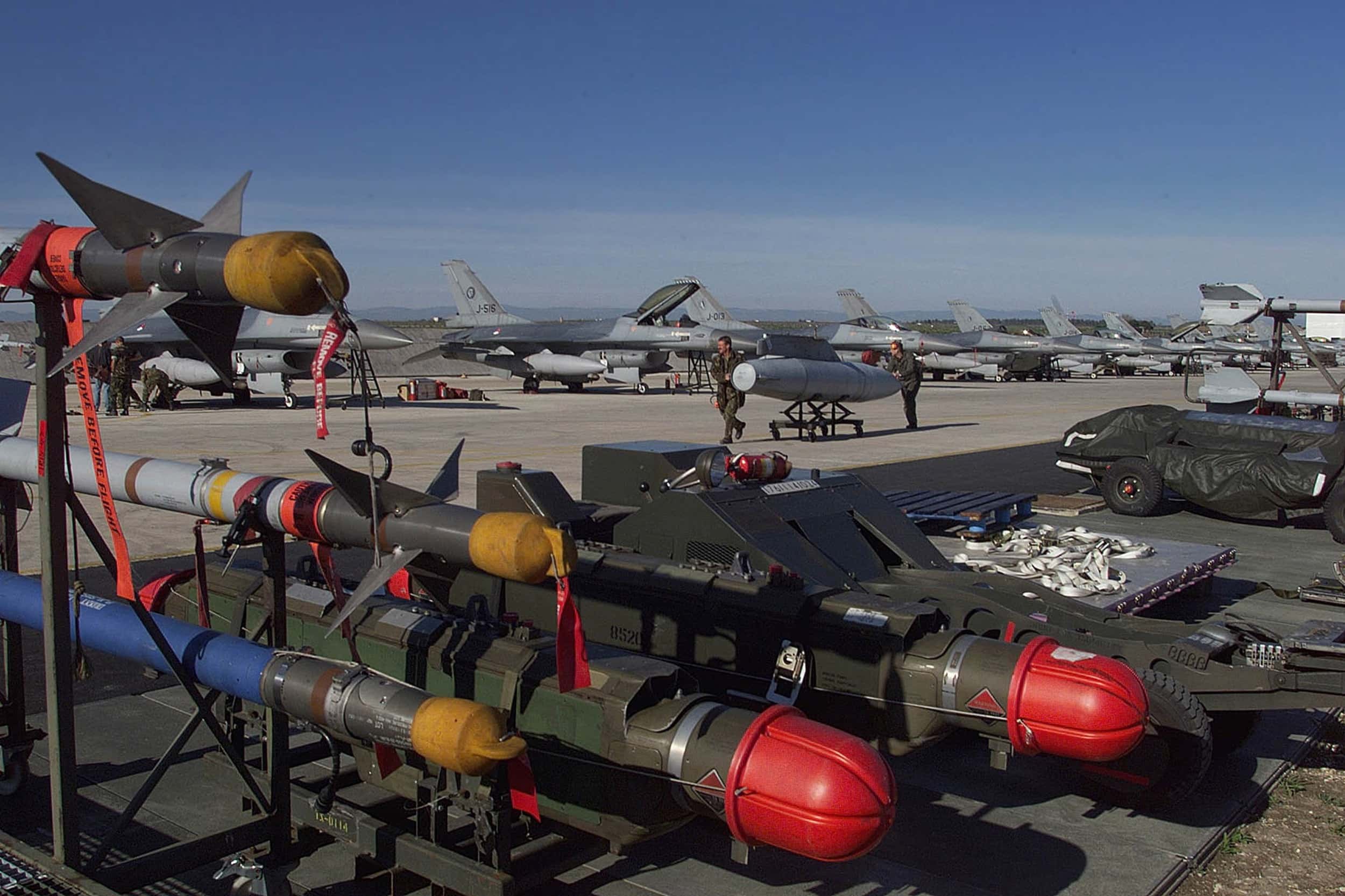 Kleurenfoto, buiten op een vliegveld. Op de voorgrond een stelling met meerdere raketten, drie lange dunne en twee dikkere. Daarnaast staat twee kleine takelwagens geparkeerd, waarvan de rechter met een zeil bedekt is. Daarachter lopen twee militairen met een F-16-brandstoftank op een rolkarretje. Verder in de achtergrond een lange rij F-16’s. De zon schijnt, er is geen wolkje aan de lucht.
