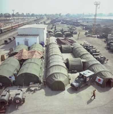 Kleurenfoto, buiten van een hoogte genomen. Meerdere groene koepeltenten die tegen en naast elkaar liggen. Op de tent en vrachtwagen rechtsvoor is een wit vlak met rood kruis aangebracht. Hier en daar lopen militairen. Rechts staan meerdere voertuigen in rijen geparkeerd, linksboven in de achtergrond een spoorlijn met trein.