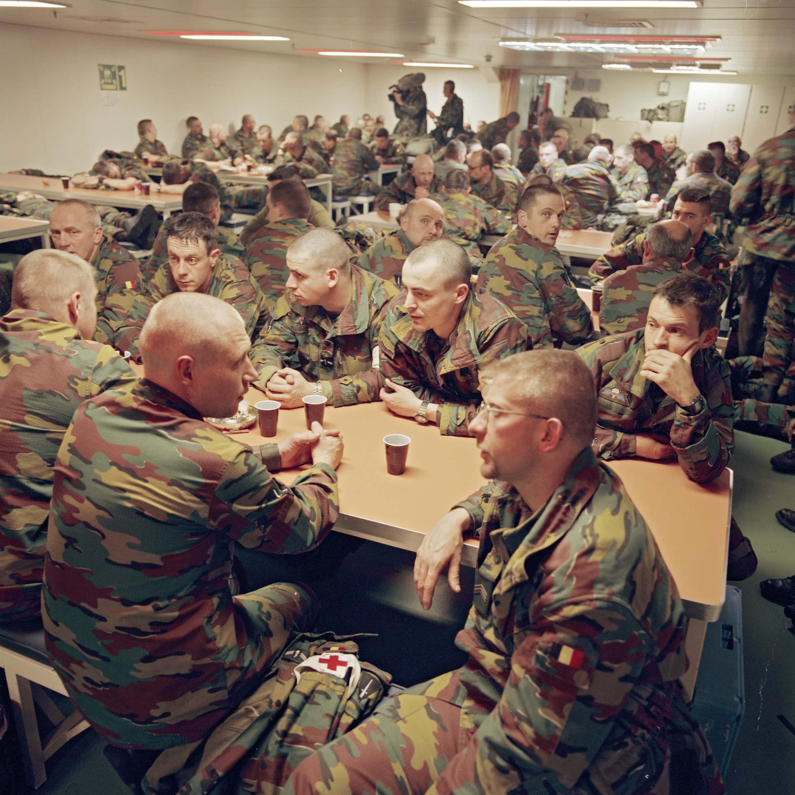 Kleurenfoto, binnen. Tientallen militairen in camouflagekleding zitten in een kantineruimte in groepen aan tafels. Velen hebben een bekertje voor zich of in de hand. Enkelen kijken in de camera, de militair rechtsvoor heeft een Belgisch vlaggetje op zijn linker mouw. In de achtergrond is een videocameraploeg opnames aan het maken.