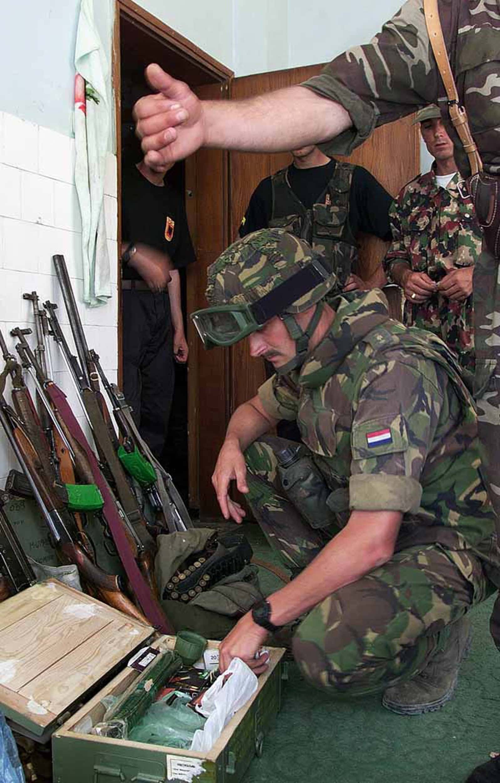 Kleurenfoto, binnen. Een militair in camouflagekleding en een helm op (grote veiligheidsbril op de helm) zit op z’n hurken naast een open kistje op de grond, op zijn linkermouw een Nederlands vlaggetje. In het kistje zitten plastic tasjes en een groene kijker. Tegen de muur staan geweren en tassen met munitie. In de achtergrond staan drie mannen naast een openstaande deur en in de deuropening.