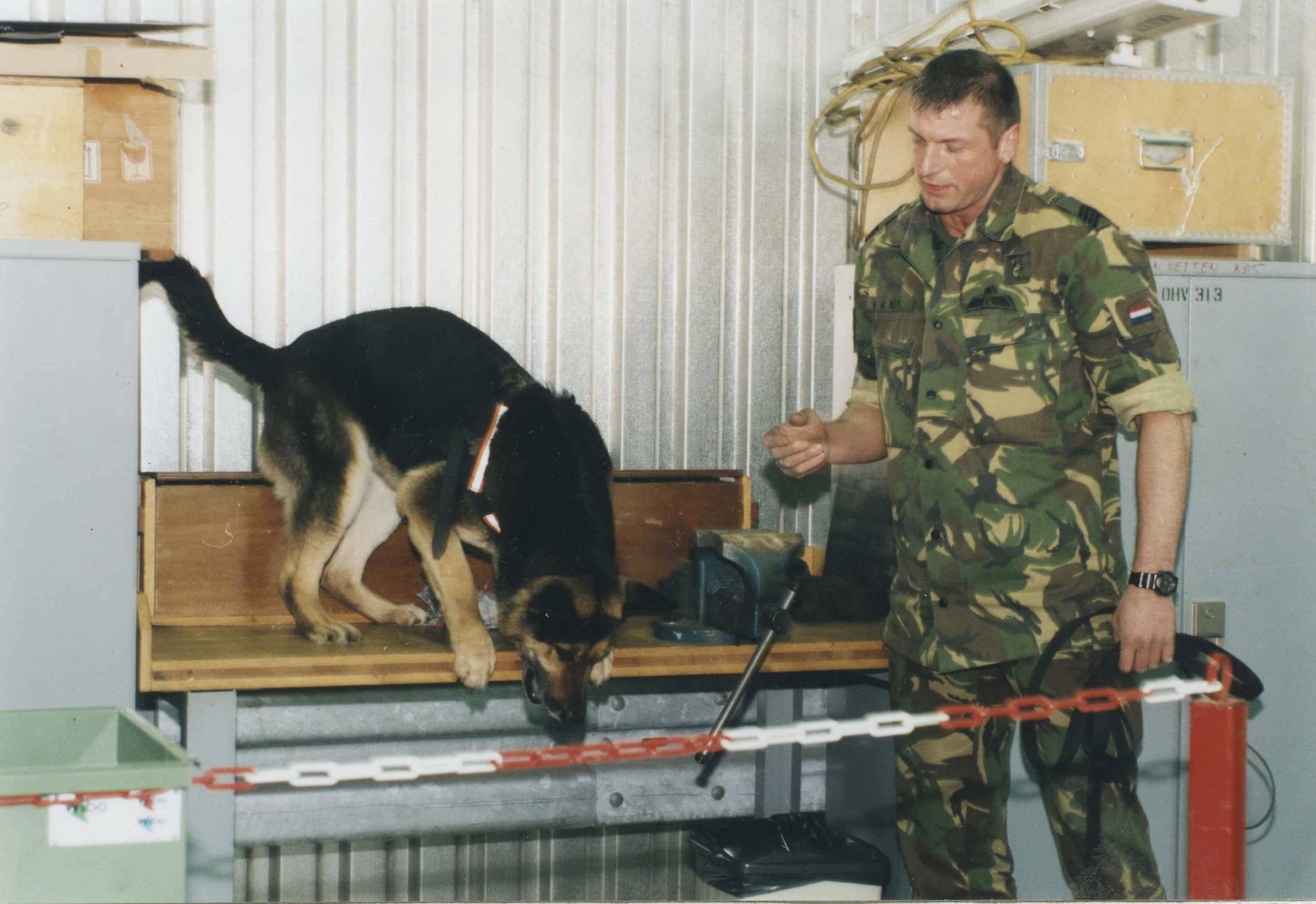 Kleurenfoto, binnen. Een hond staat op het punt om van een werktafel te springen, rechts haar baasje in camouflagekleding en de hondenlijn in zijn linkerhand. Tussen hen in, op de tafel, een bankschroef. Links en rechts van de werktafel kasten met kosten bovenop, daarachter een muur van staalplaten. Voor de hond en het baasje een rood-witte veiligheidsketting, dat rechts aan een rood paaltje vastzit.