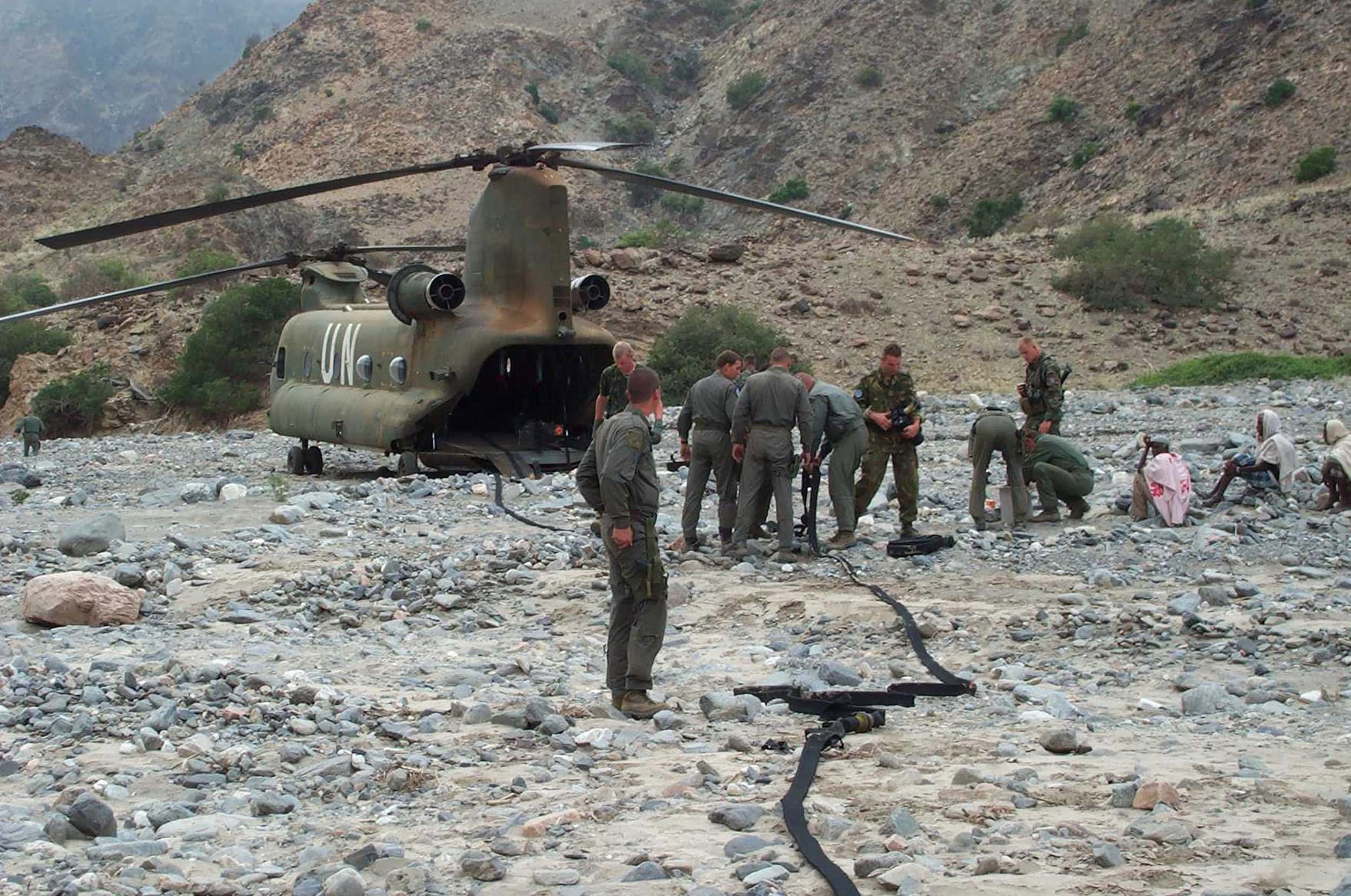 Kleurenfoto, in een rotsige omgeving staat een grote helikopter aan de grond, rotorbladen voor- en achterop, met open achterkant, er loopt een pompslang naar buiten, deze loop richting de cameraman en verder buiten beeld. In het midden van de foto is een groep van negen militairen met de slangen in de weer. Helemaal rechts zitten drie Afrikaanse mannen op de grond.
