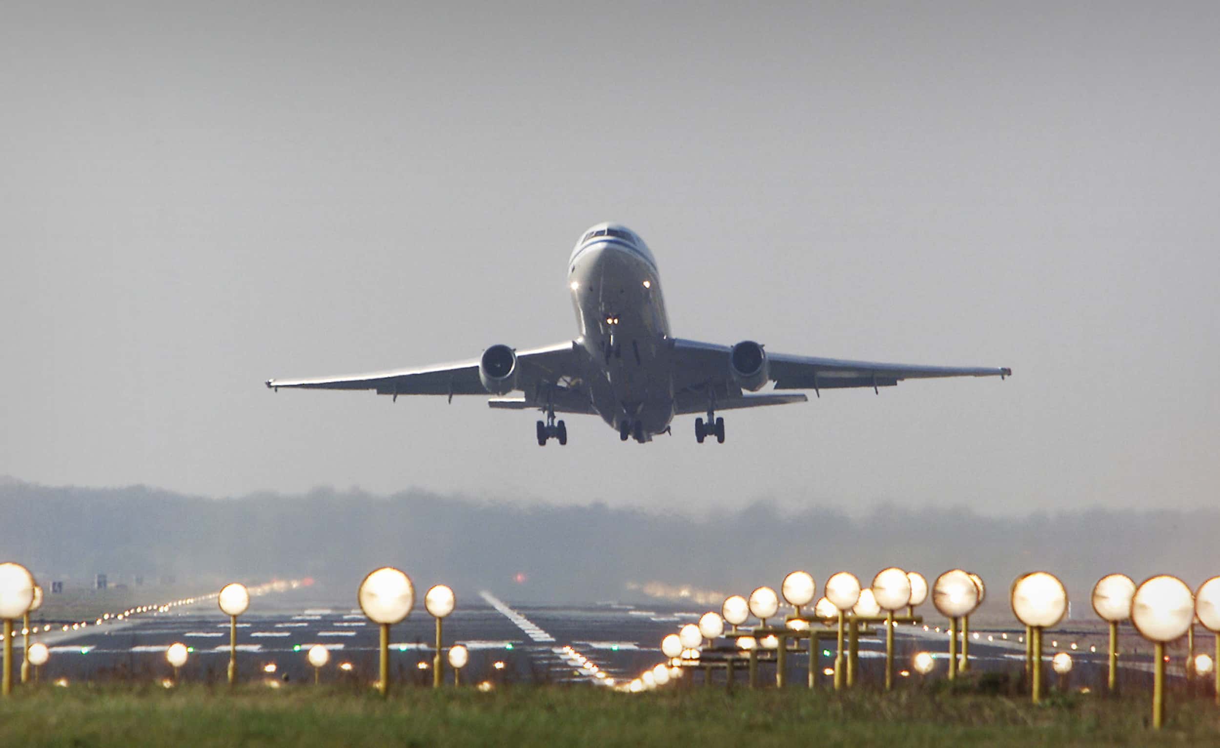 Kleurenfoto, buiten. Een vliegtuig stijgt op en hangt boven de startbaan, op de voorgrond tientallen lampen op een rij in het verlengde van de baan. In de achtergrond bomen, te zien door de warme lucht die uit het vliegtuig komt.