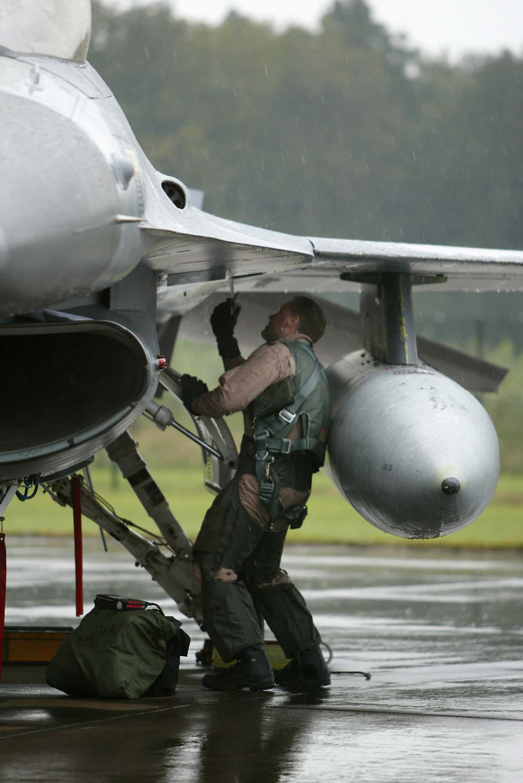 Kleurenfoto, buiten op een vliegveld. Een militair staat onder de vleugel van een F-16, die voor de helft te zien is, en houdt met zijn rechterhand iets vast, waarmee hij in de vleugel werkt. Voor zijn voeten ligt een stoffen tas. Het regent.