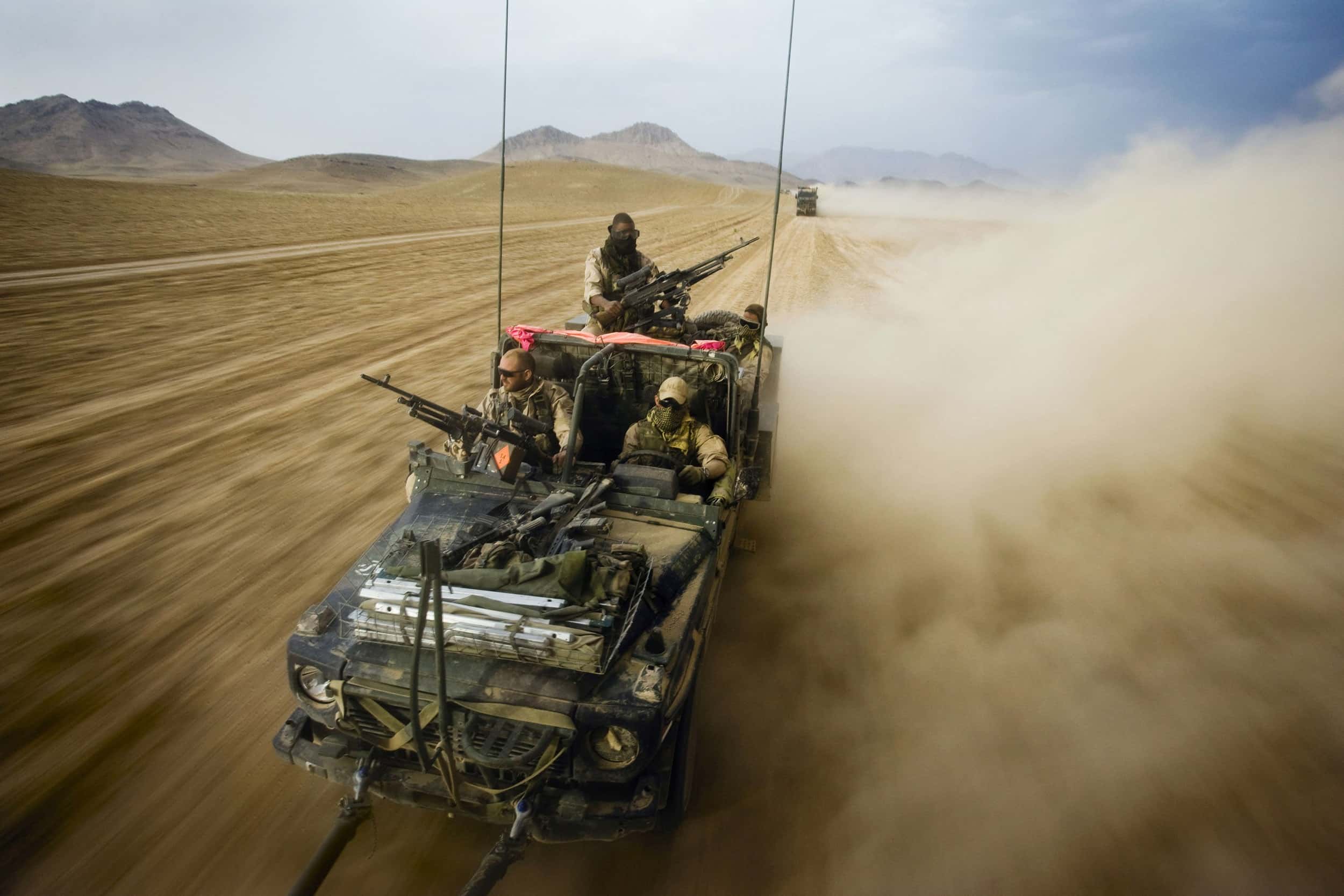 Kleurenfoto. Vier militairen zitten in een jeep, ze dragen camouflagekleding, zonnebrillen en doeken voor hun neus en mond. Voor- en achterop de jeep zijn mitrailleurs gemonteerd, aan de zijkanten steken twee lange antennes omhoog. Ze rijden door een woestijn en veroorzaken een grote stofwolk. Achter hen een ander militair voertuig.