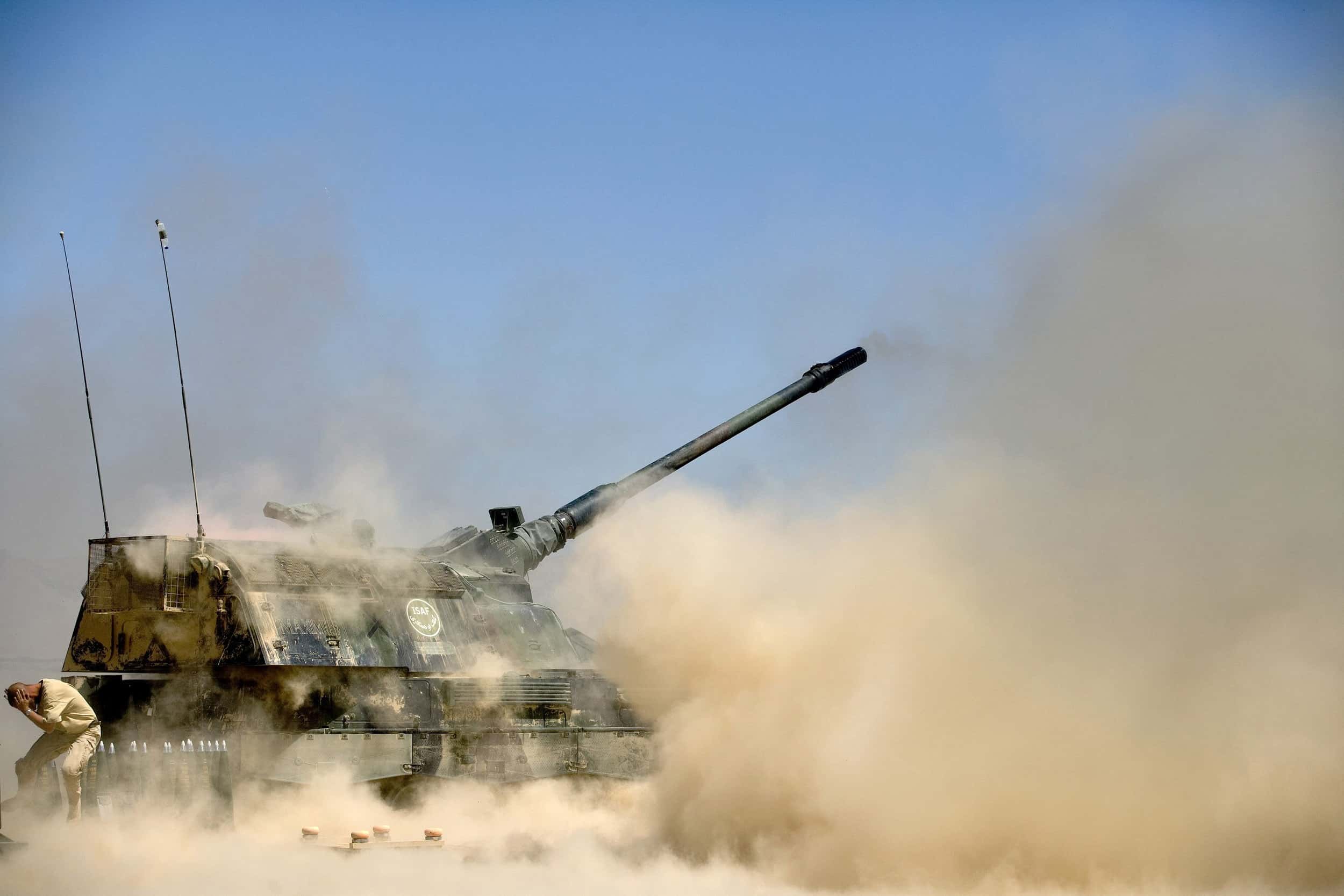 Kleurenfoto. Een groot kanon heeft net afgevuurd. Overal hangt stof en zand in de lucht. Links staat een militair met zijn rug naar het kanon voorovergebogen, hij bedekt zijn oren.