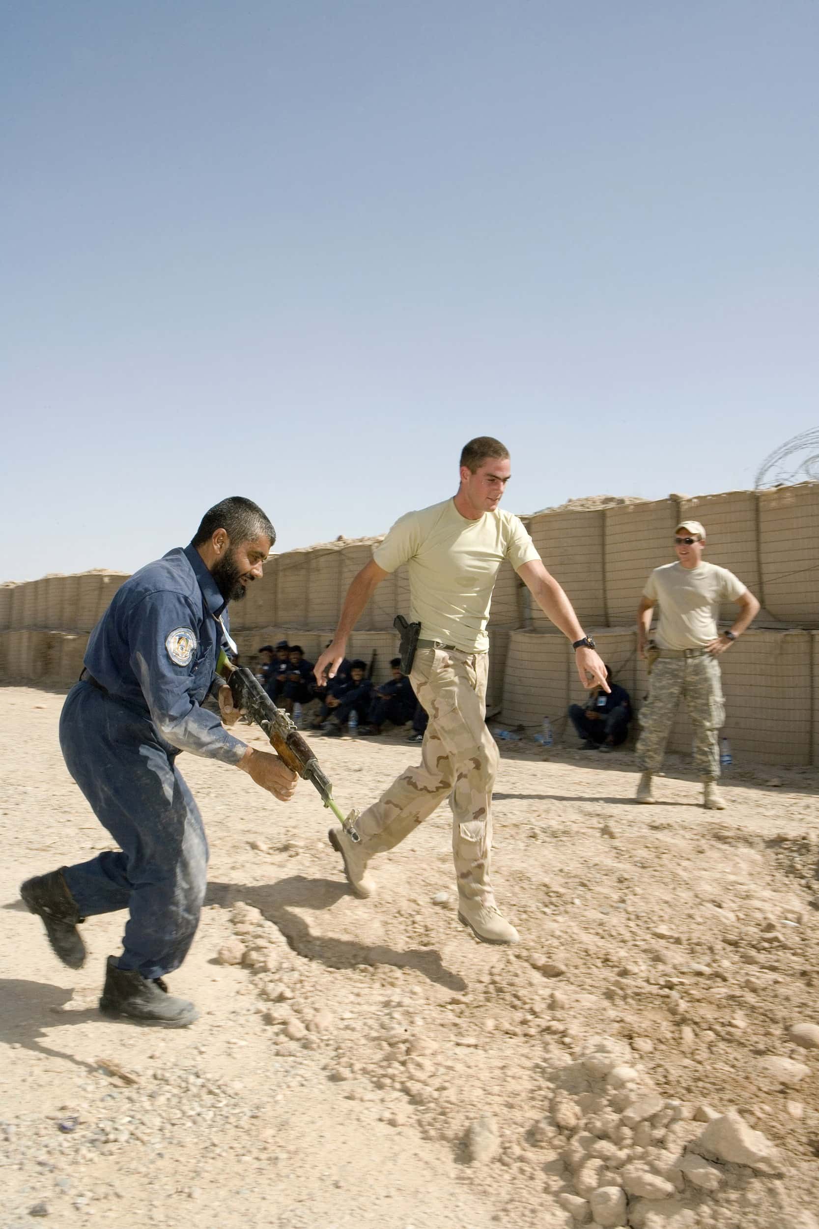 Kleurenfoto. Twee militairen lopen buiten. De linker draagt een blauwe overall, heeft een baard en houdt een geweer vast. De rechter draagt beige kleding, een pistool aan de riem en hij wijst naar de grond. In de achtergrond staat een andere militair met zijn handen in zijn zij, ook in beige kleding. Hij draagt een pet en zonnebril. In de schaduw van een muur zit een rij mannen in blauwe overalls.