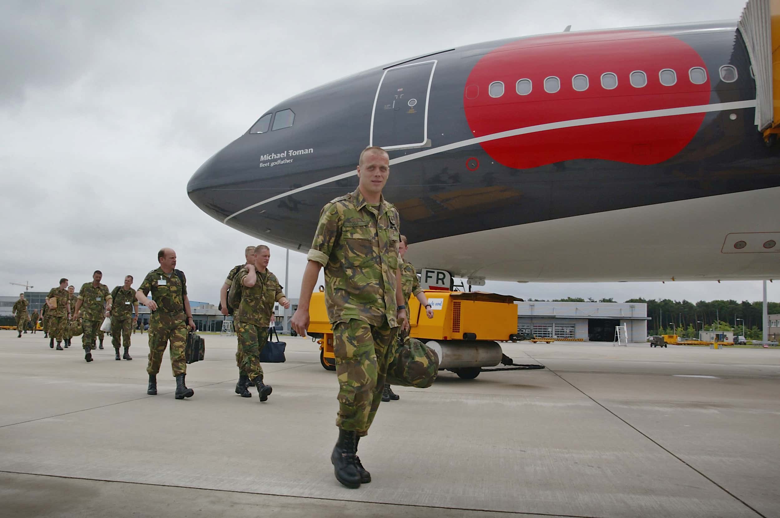 Kleurenfoto, buiten op een vliegveld. Militairen in camouflagekleding lopen vanuit links achter elkaar in een rij, ze dragen allemaal kleine tassen in hun hand of op hun rug. Vanaf rechts torent een groot vliegtuig boven hen uit, onder de neus daarvan een oranje pushback truck.