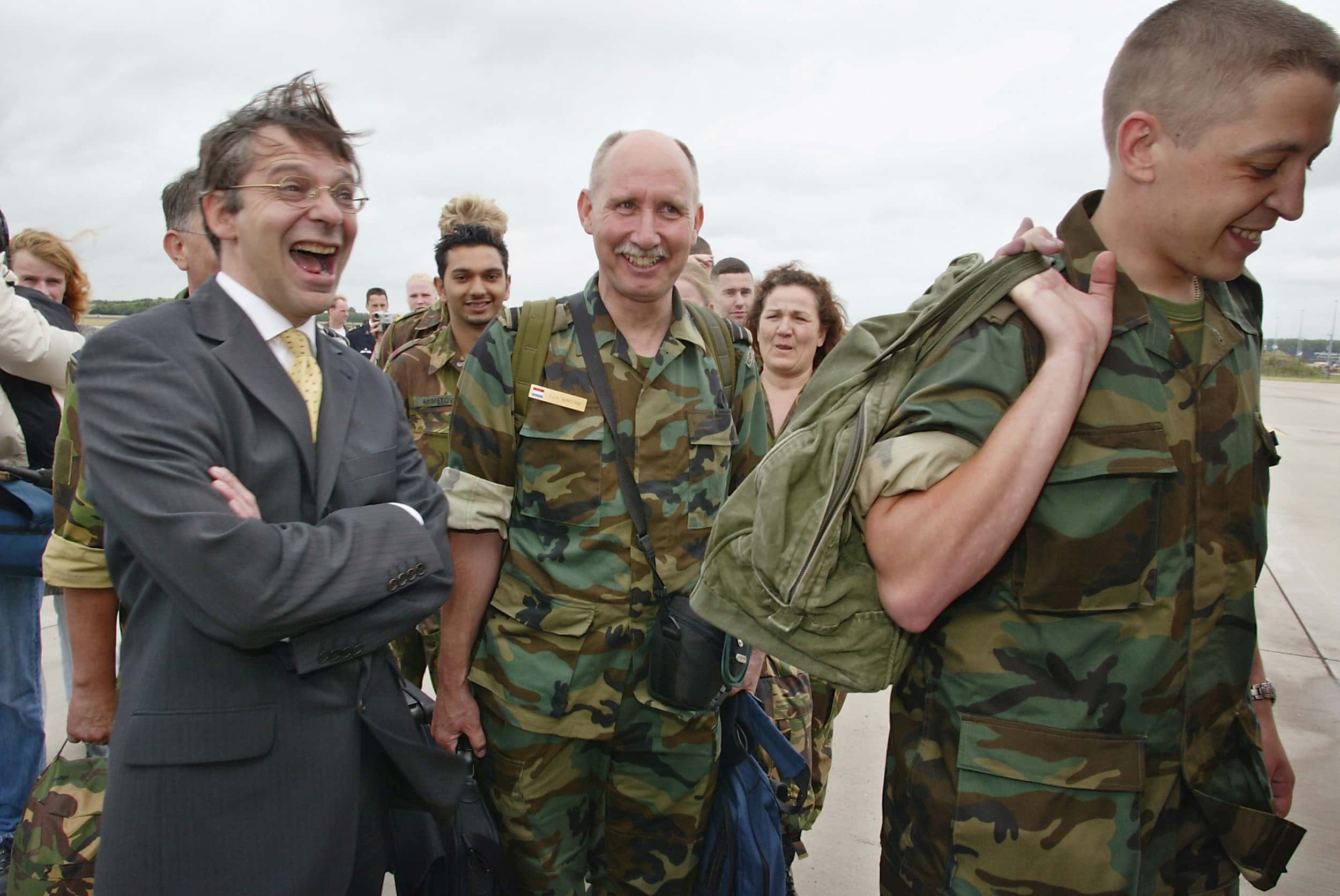 Kleurenfoto, buiten. Militairen lopen in lachend en glimlachend een rij langs andere mensen, zij dragen camouflagekleding en hebben een tas in hun hand of op hun rug. Links een man in pak met stropdas, hij staat met zijn armen over elkaar en lacht hardop. In de achtergrond mannen en vrouwen in burgerkleding.