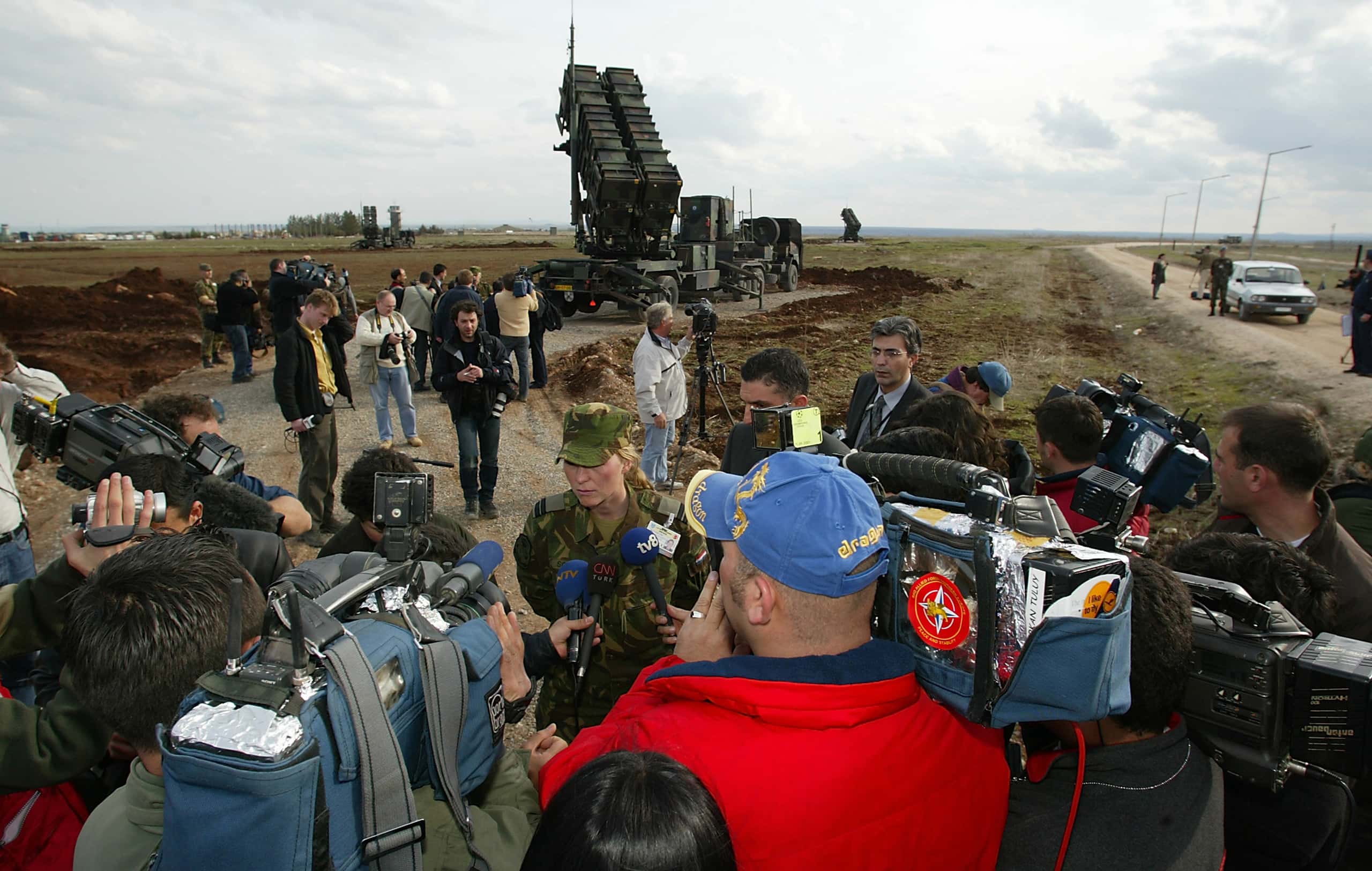 Kleurenfoto. In de achtergrond een vrachtwagen met twee grote langwerpige kisten op de achterlader, die schuin omhoog gericht staat. Op de voorgrond staat een vrouwelijke militair de pers te woord. Vijf cameramannen hebben hun camera op haar gericht, drie microfoons worden voor haar gehouden. Tussen de militair en de vrachtwagen staan fotografen die foto’s maken van de wagen en de omgeving.