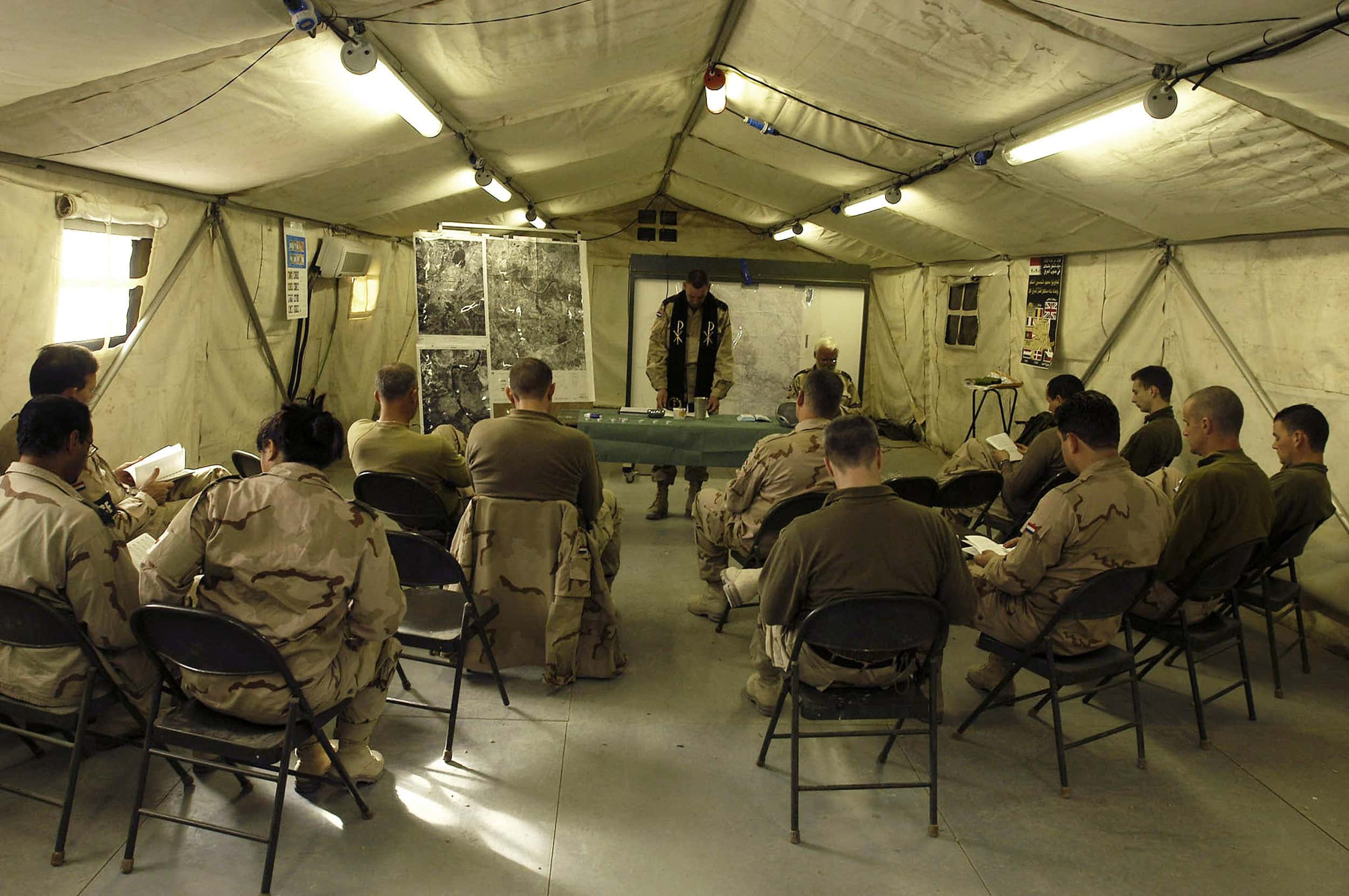 Kleurenfoto. Twaalf militairen zitten (vanaf de rug gezien) op stoelen in een tent, in het midden tussen de stoelen loop een klein gangpad. Ze lezen in een boekje. Vooraan staat een gedekte tafel, daarachter een militair met een stola om de schouders. Ook hij leest uit een boekje dat voor hem op tafel ligt. Naast hem een groot bord met kaarten en foto’s. Aan het plafond van de tent hangen tl-buizen.