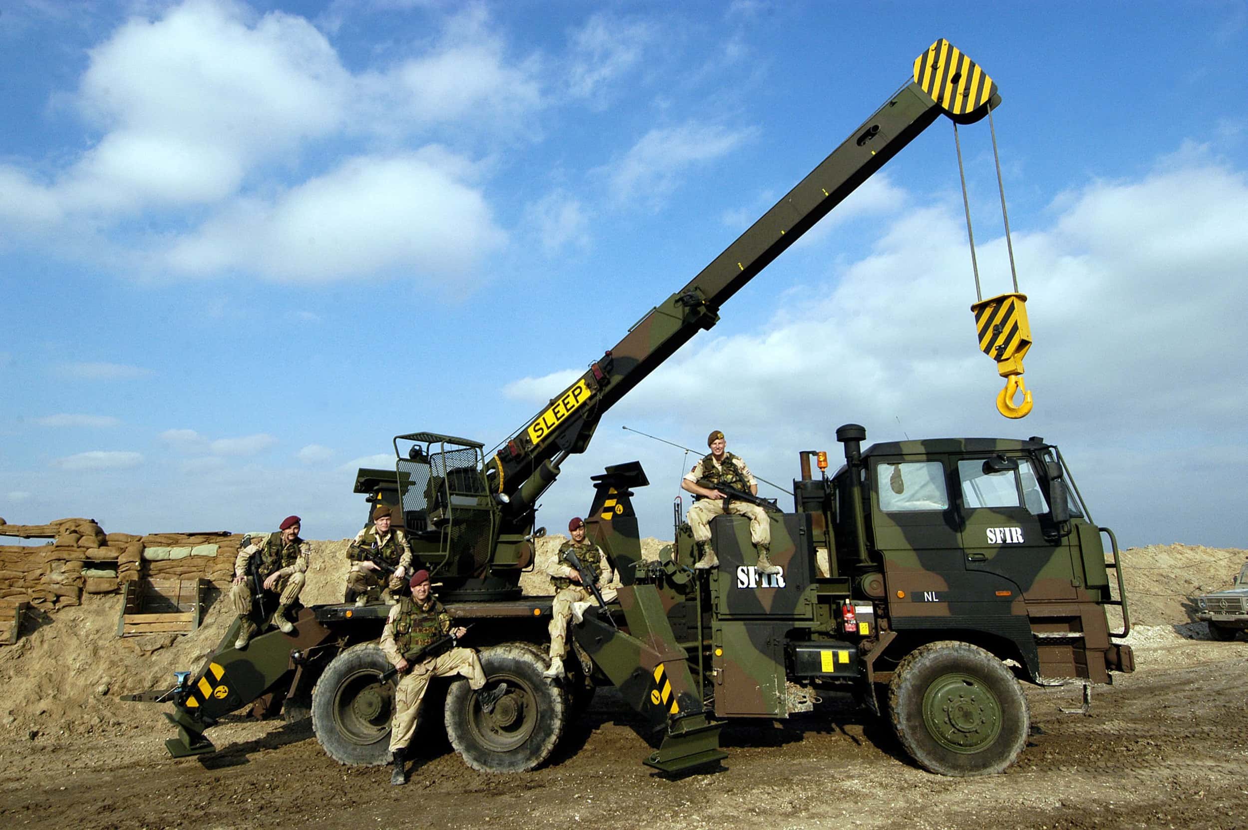 Kleurenfoto. Te zien is een grote wagen die ergens geparkeerd staat. De wagen is in camouflagekleuren geschilderd, op de zijkant staat twee keer in het wit SFIR geschreven. Vanaf de achterkant steekt een lange arm omhoog, met aan het einde daarvan een grote haak. Op de wagen zitten vijf militairen, ze dragen rode baretten en een wagen.