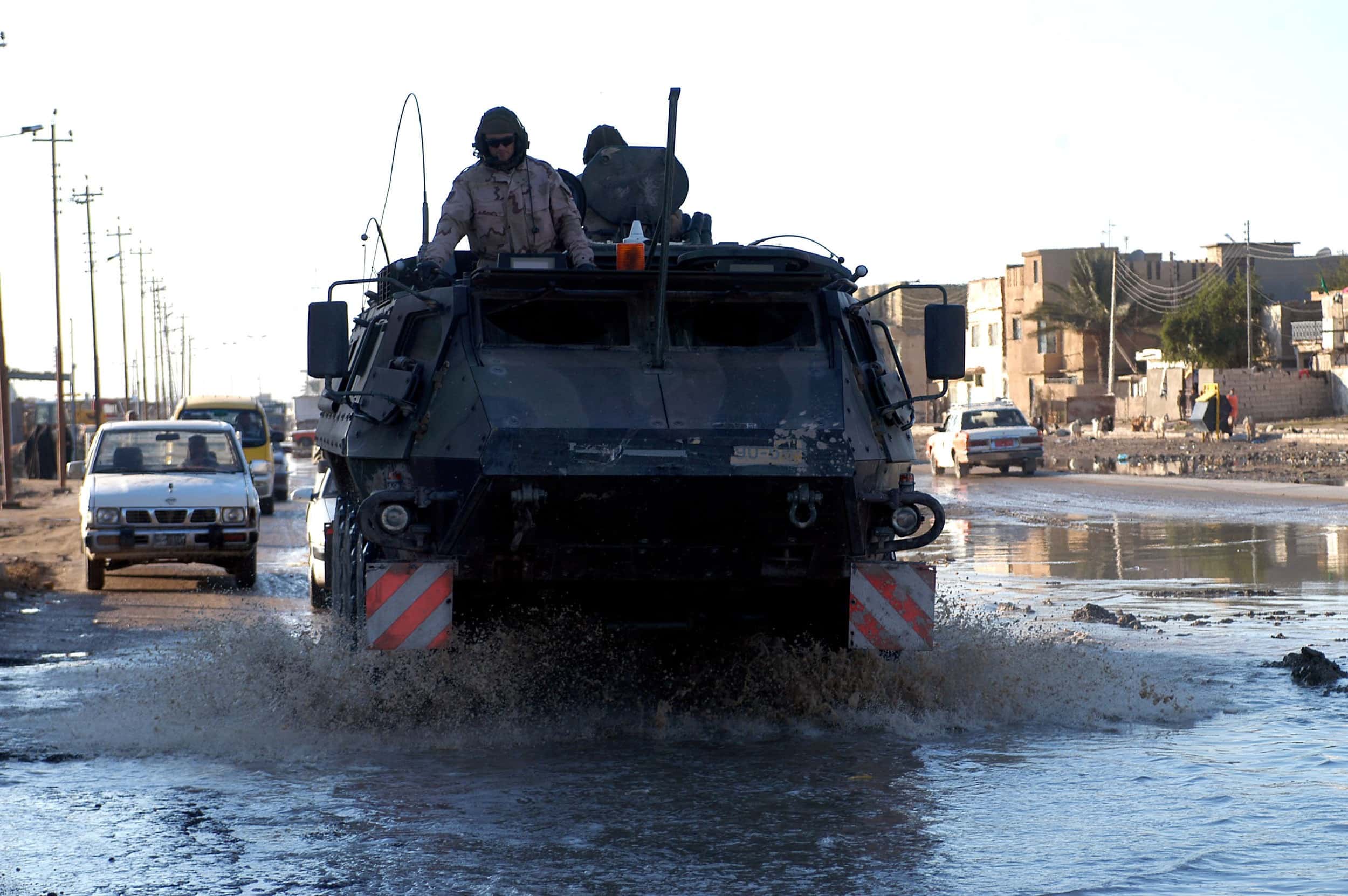 Kleurenfoto. Een pantserwagen rijdt over straat door een grote plas water. Water spat rond de voorwielen omhoog. Vlak voor de wielen zijn twee rood-wit gestreepte waarschuwingsbordjes aangebracht. Een militair steekt aan de bovenkant van de pantserwagen met zijn bovenlichaam uit een luik. In de achtergrond auto’s en gebouwen.