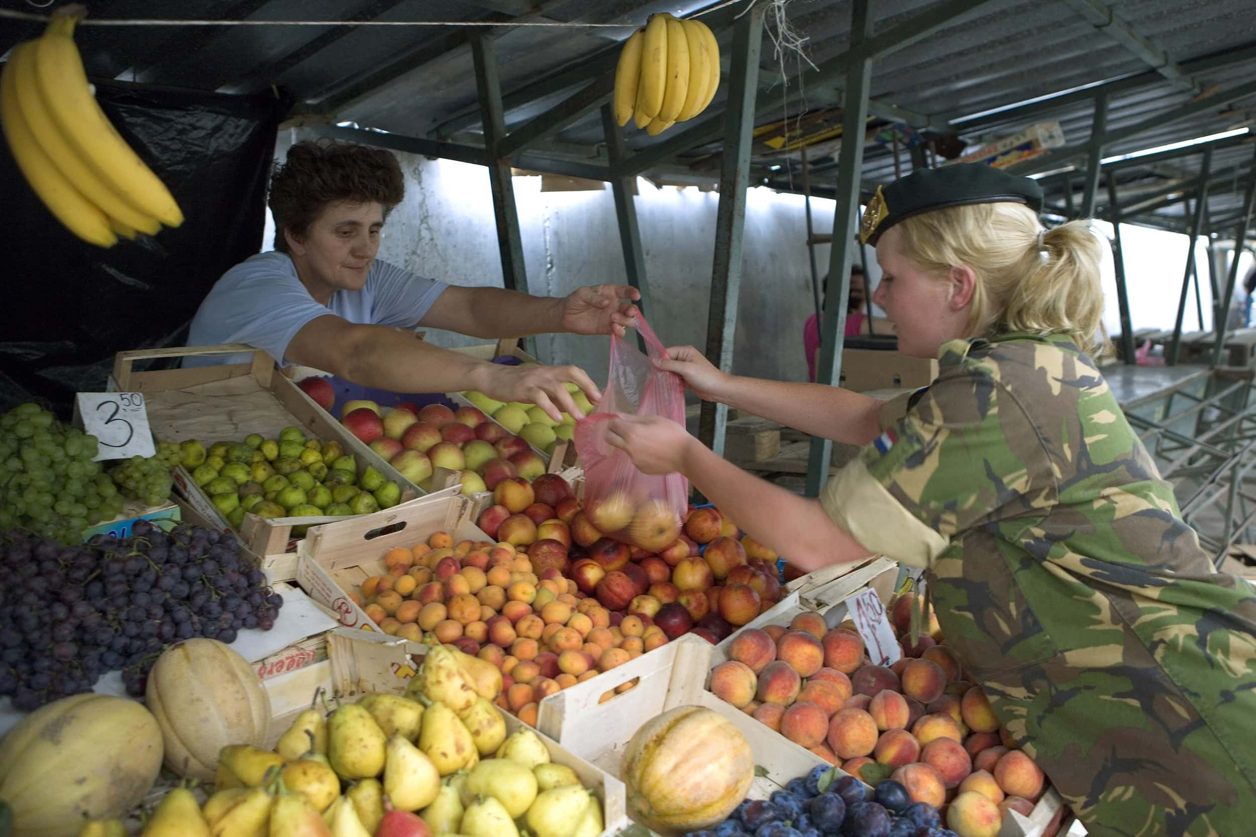 Kleurenfoto, twee vrouwen op de markt, de rechter draagt groene camouflagekleding en een baret, de linker is in burgerkleding. De vrouw links reikt een plastic zak appels aan en neemt tegelijkertijd geld in ontvangt. Ze rekken zich beiden uit boven fruit, zoals appels, perziken, peren en druiven.