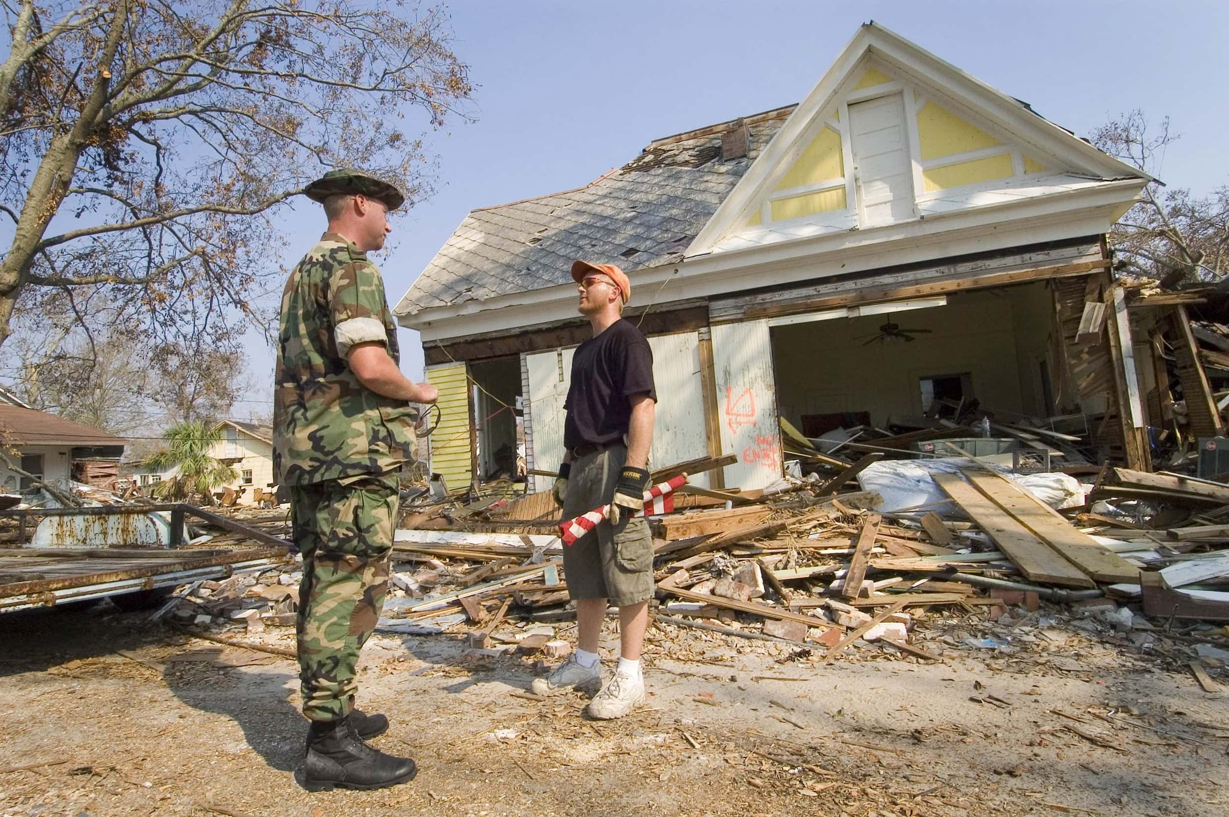 Kleurenfoto, buiten. Links staat een militair in camouflagekleding, hij draagt een zonnehoed. rechts een man in burgerkleding. Hij draagt een korte broek, tuinhandschoenen, een petje en een zonnebril, in zijn hand een opgerolde rood-witte vlag. Achter hen een zwaar beschadigd huis met opengeslagen garage-gat en heel veel puin op de grond.