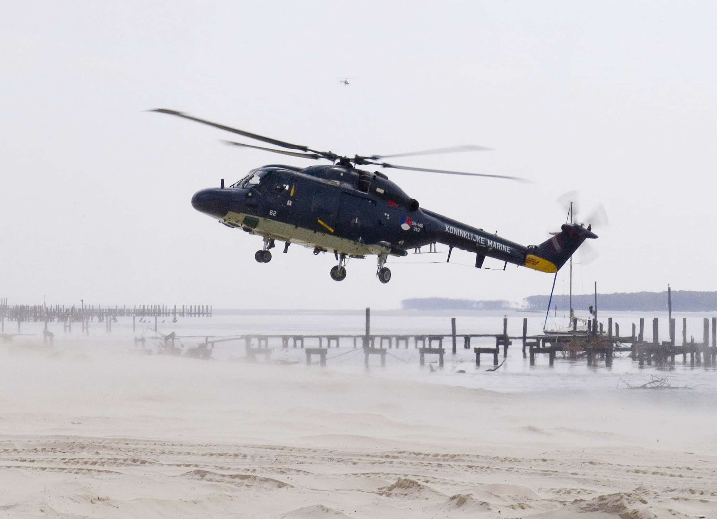 Kleurenfoto, buiten op een strand. Een helikopter hangt in de lucht, daaronder stuift zand weg. Op de staart van het toestel staat Koninklijke Marine. In het zand zijn sporen van autobanden, op de achtergrond beschadigde houten pieren. De lucht is grijs.