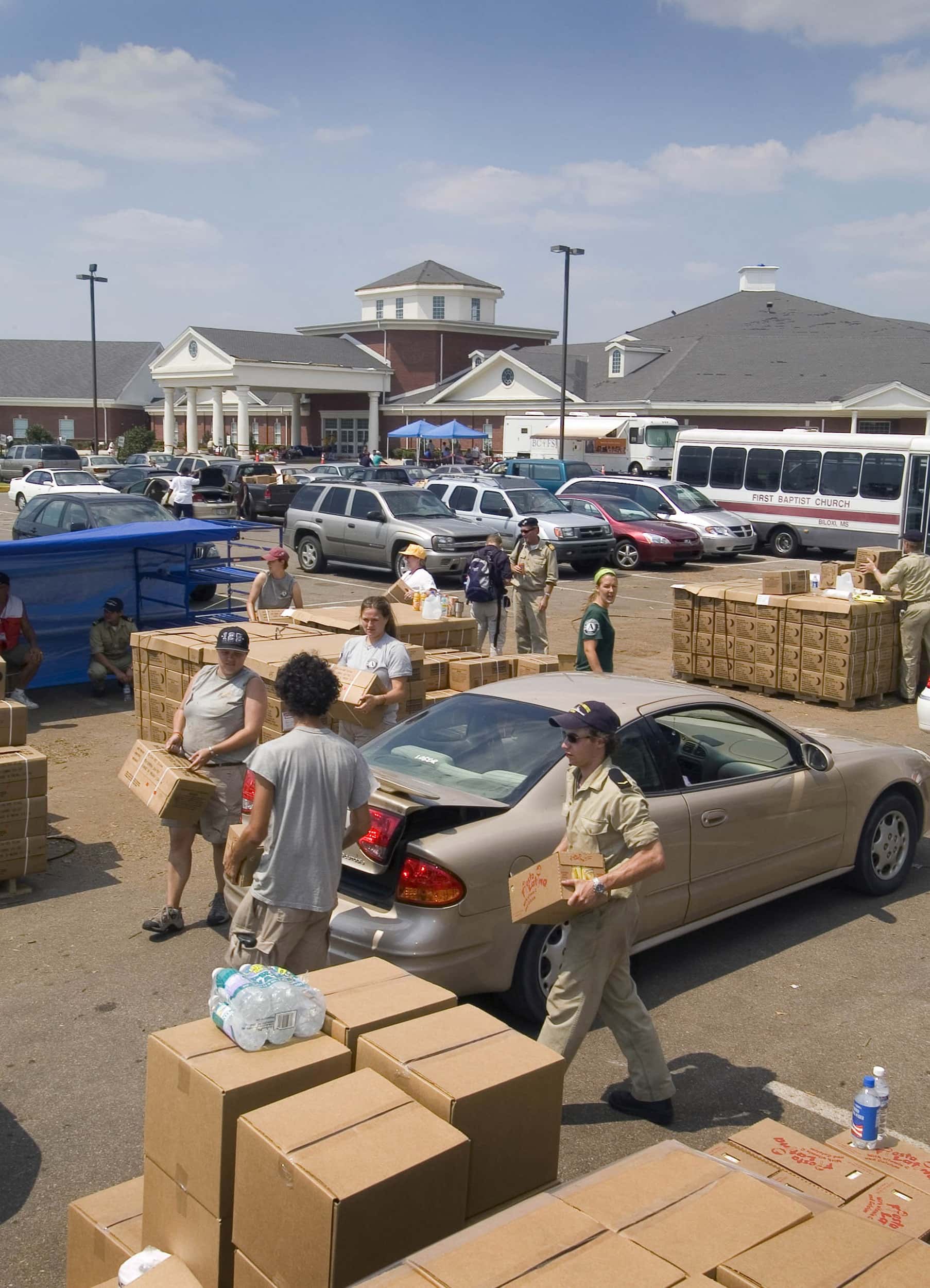Kleurenfoto, buiten op een parkeerplaats. Vier mensen, waaronder een militair met petje en zonnebril, dragen kartonnen dozen naar de achterkant van een auto, de achterbak staat half open. Om hen heen meerdere stapels dozen, waar tussen andere mensen lopen en zitten. In de achtergrond staan veel auto’s en een bus geparkeerd. Daarachter een groot winkelcentrum.