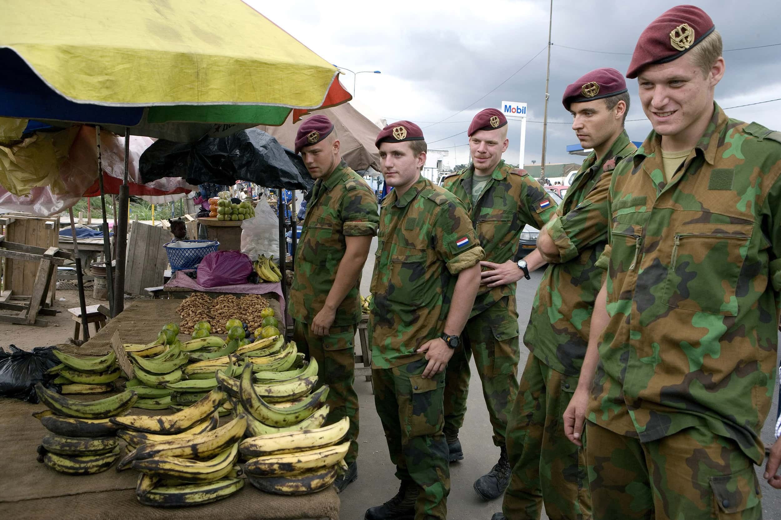 Kleurenfoto, vijf militairen met rode baretten en camouflagekleding staan bij een tafel met bananen.
