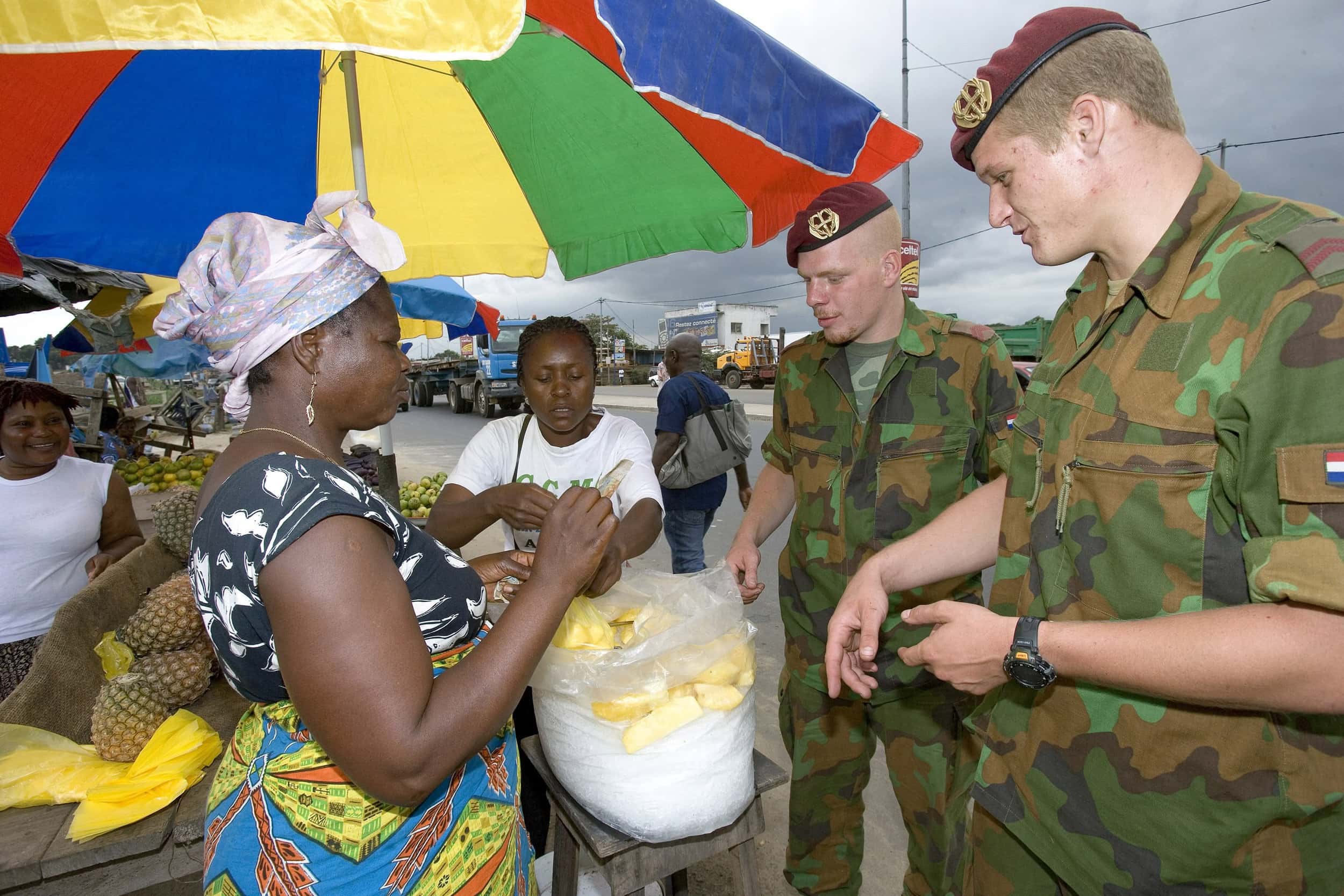 Kleurenfoto, rechts staan twee militairen in camouflagekleding, links drie marktvrouwen onder een gekleurde parasol, tussen hen in een zak met stukken ananas.
